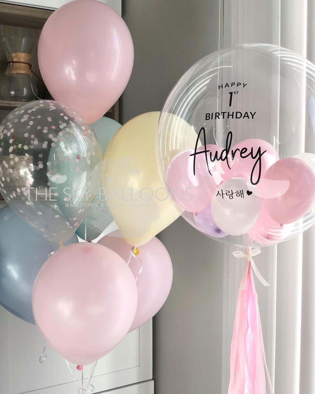 A bouquet of personalized balloons in shades of pink, blue, and transparent, with 'Happy Birthday' and a name 'Audrey' printed on a pink balloon, along with other balloons with different messages.