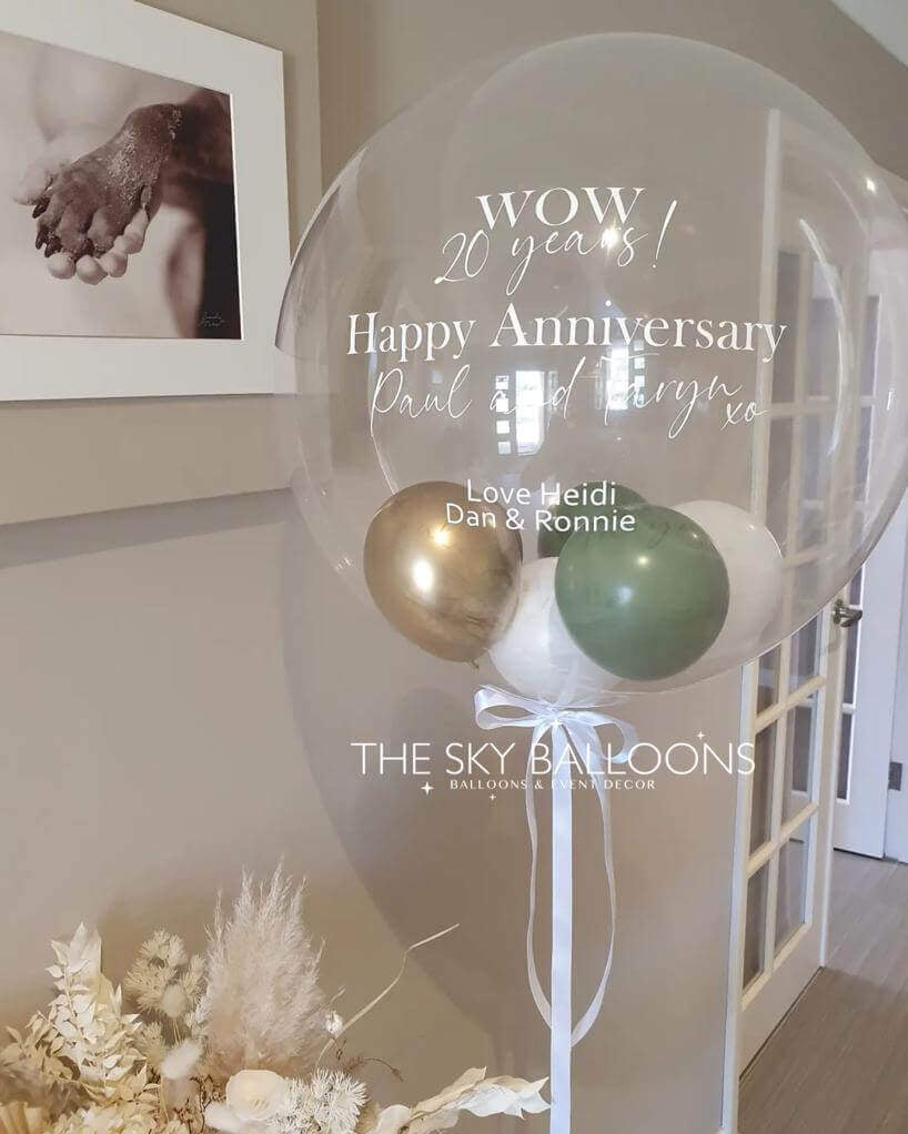 A clear round balloon with gold, white, and green tassels, featuring a celebratory message for a happy anniversary, displayed in a home environment.