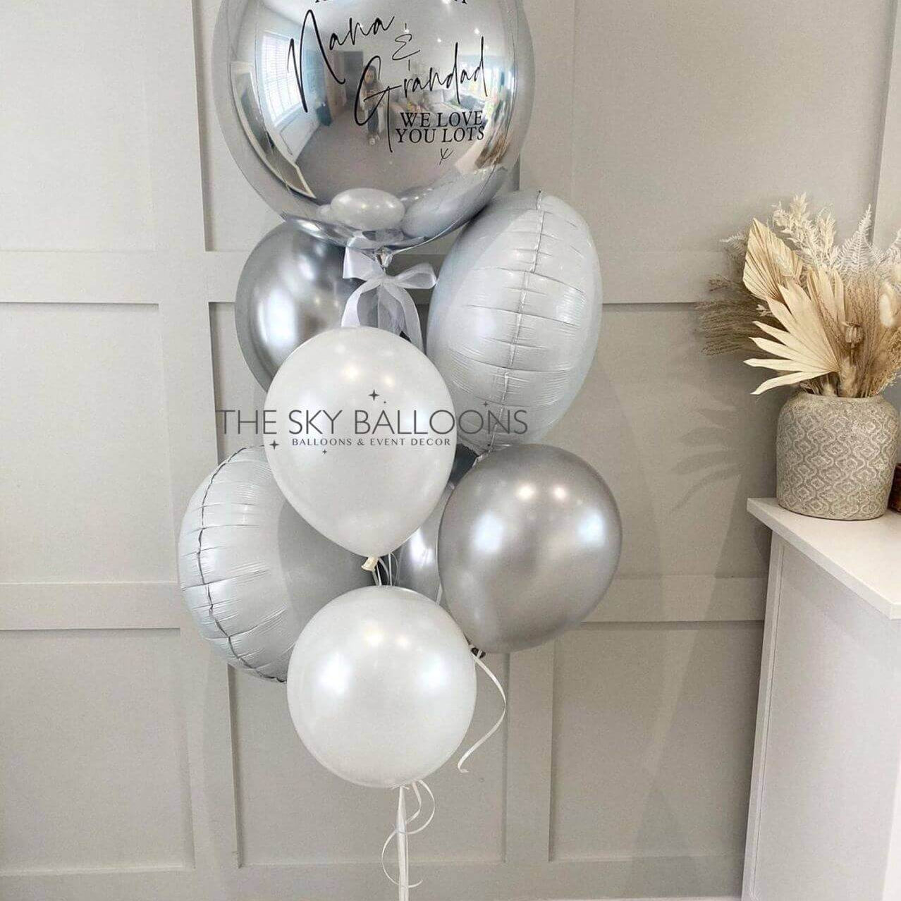 A bouquet of latex balloons in various sizes, including a large customized orb, with a silver foil finish. The balloons are tied with ribbons and have a celebratory message printed on the orb.