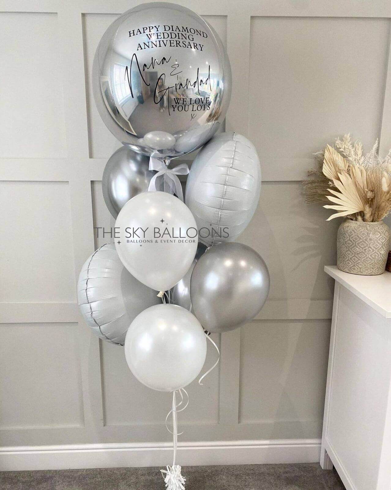 A bouquet of latex balloons in various sizes, including a large customized orb, with a silver foil finish. The balloons are tied with ribbons and have a celebratory message printed on the orb.