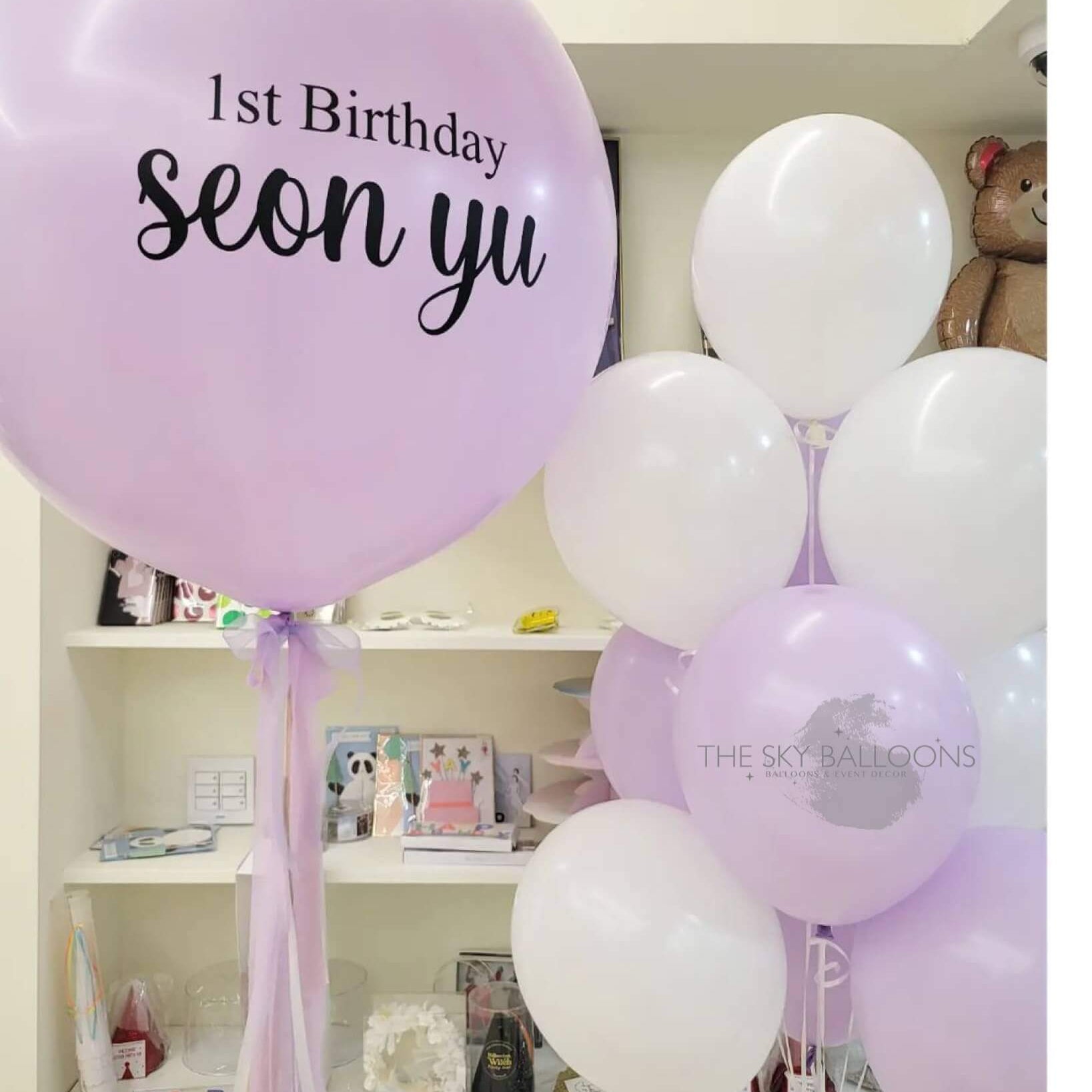 A bouquet of pink and white balloons with one of them customized with the text '1st Birthday Seony'. The balloons are tied with ribbons and placed in a shop display.