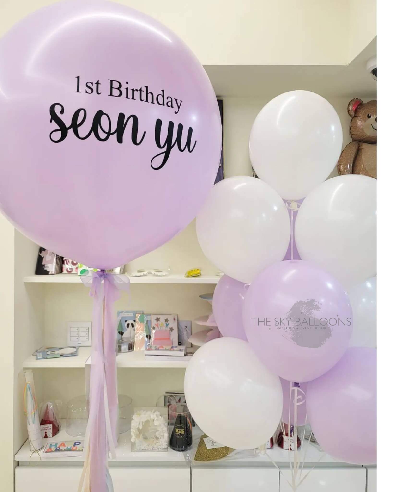 A bouquet of pink and white balloons with one of them customized with the text '1st Birthday Seony'. The balloons are tied with ribbons and placed in a shop display.
