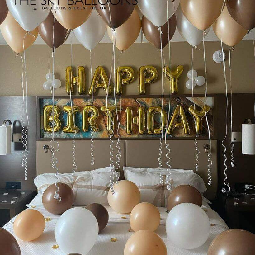 A set of balloons arranged for a birthday celebration, with a 'Happy Birthday' banner, in a room with ceiling and floor balloons in shades of gold, silver, white, and bronze.