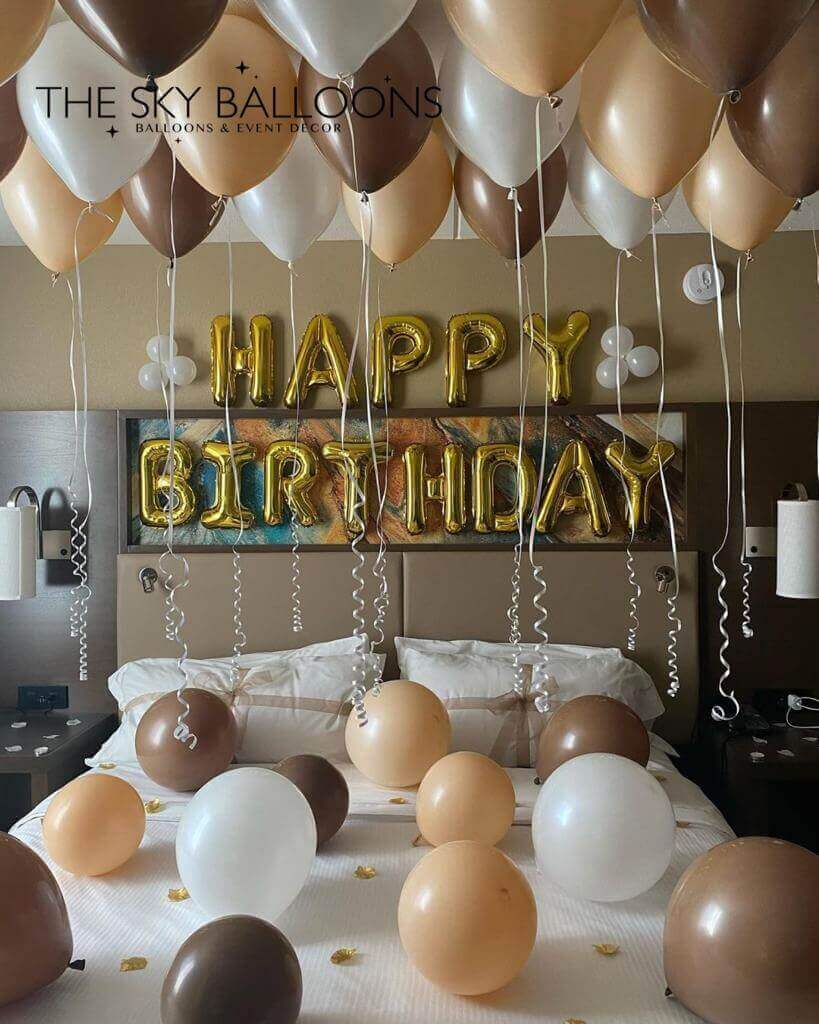 A set of balloons arranged for a birthday celebration, with a 'Happy Birthday' banner, in a room with ceiling and floor balloons in shades of gold, silver, white, and bronze.