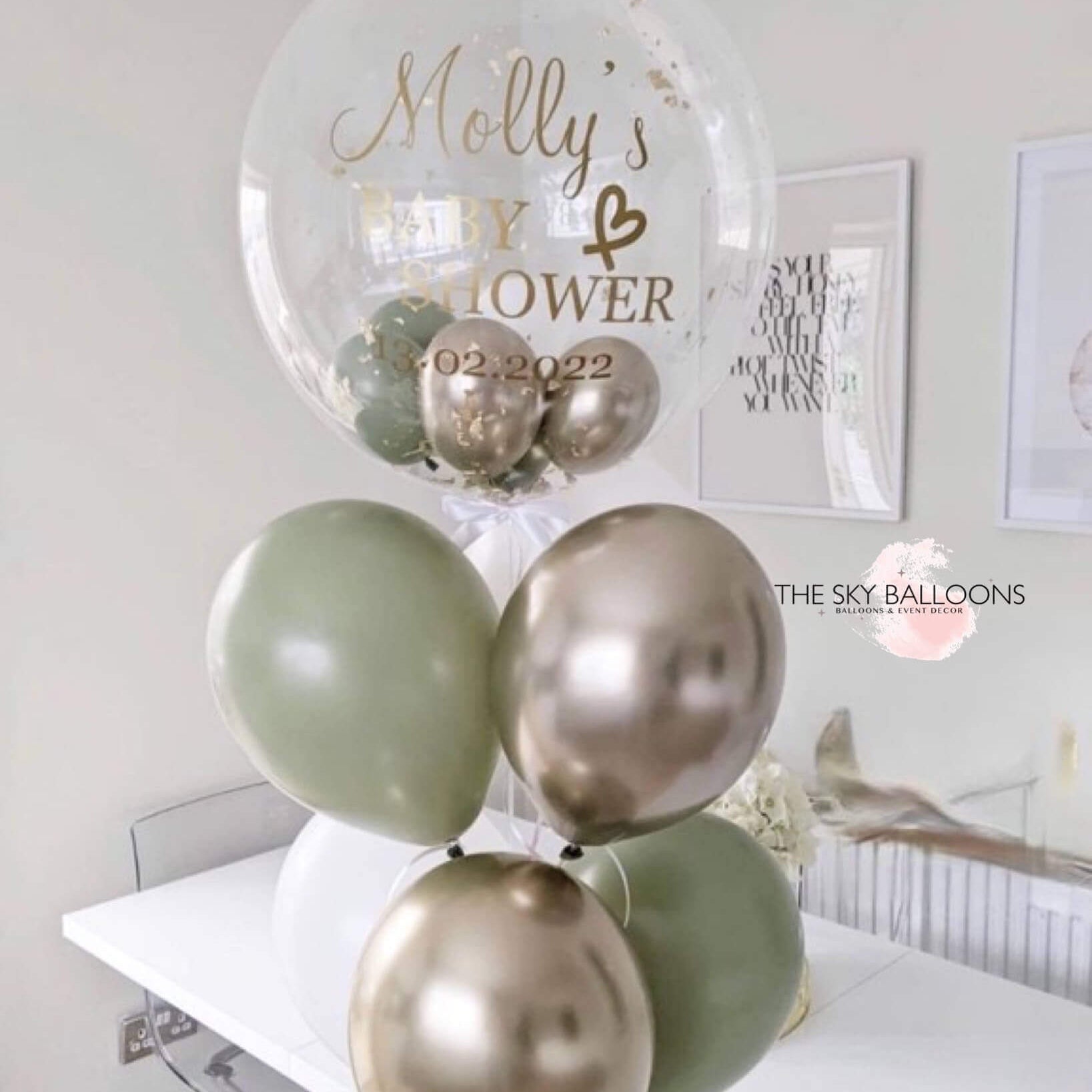 A bouquet of balloons including eucalyptus, white, and chrome gold colors, with a personalized jumbo balloon that reads 'Molly's Baby & Hair' with a date.