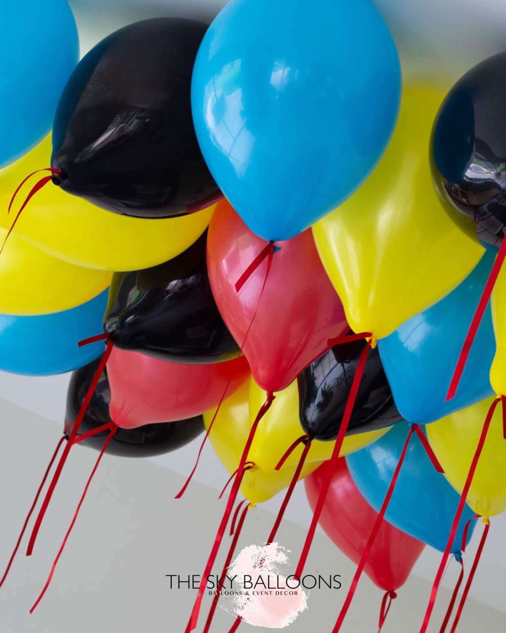 A cluster of inflated ceiling balloons in red, blue, yellow, and black colors with ribbons attached, delivered by The Sky Balloons.
