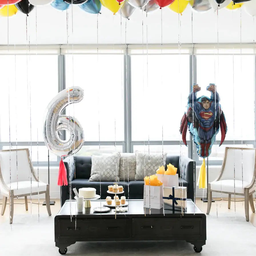 A room decorated with superhero-themed balloons including a jumbo number 6 balloon, superhero character balloons in red, blue, and silver colors, and tassels. The balloons are displayed above a table with birthday cake and decorations.