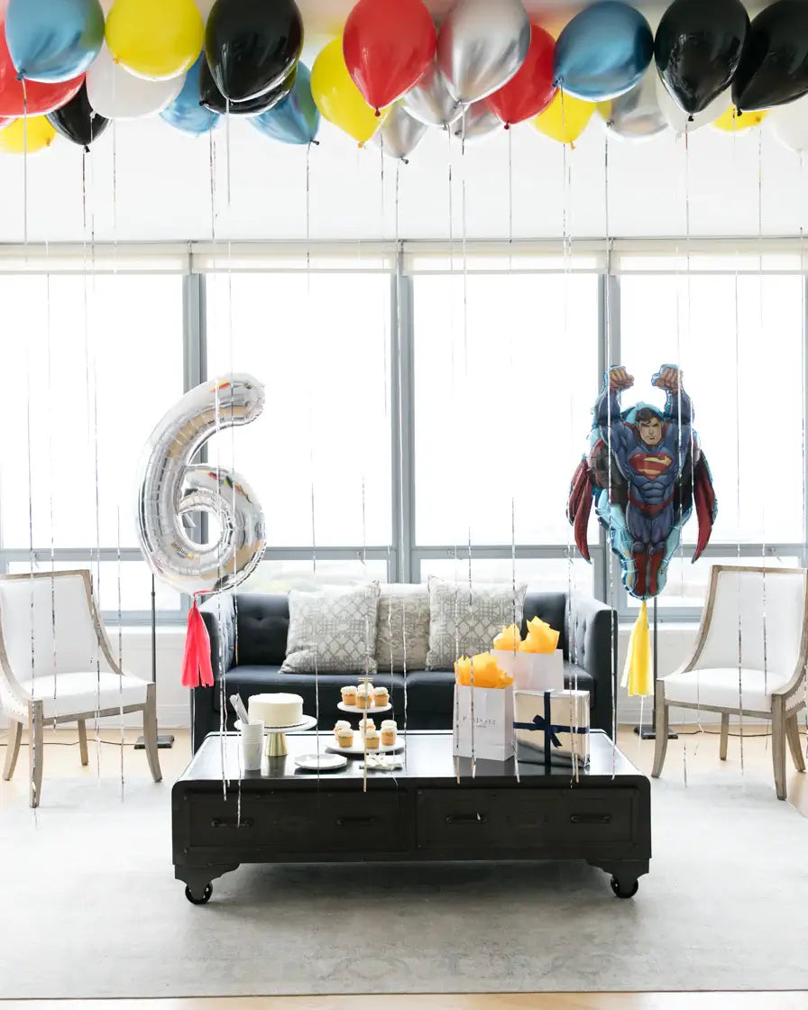 A room decorated with superhero-themed balloons including a jumbo number 6 balloon, superhero character balloons in red, blue, and silver colors, and tassels. The balloons are displayed above a table with birthday cake and decorations.