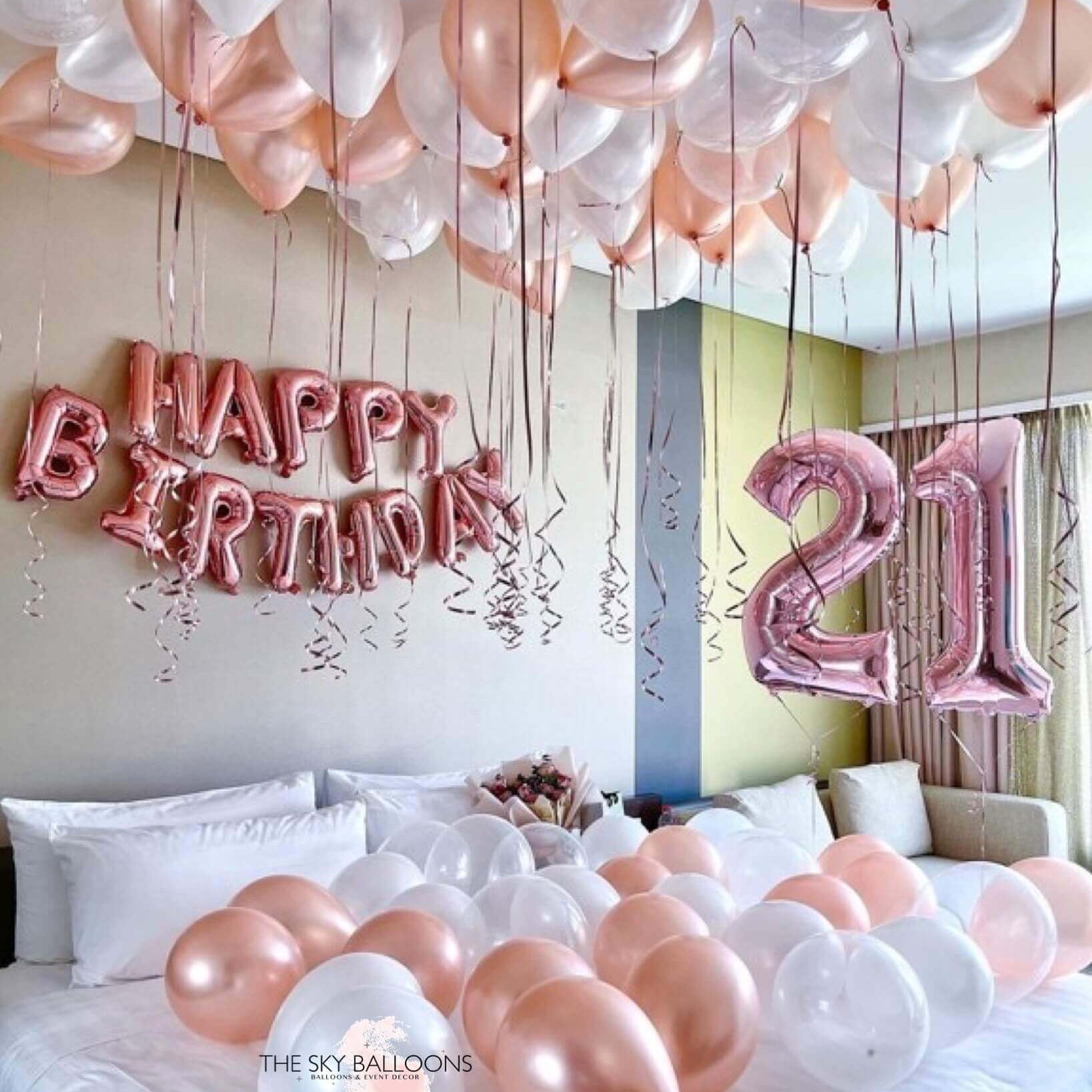 A room decorated with rose gold and pearl white balloons, including a 'Happy Birthday' banner and large numbered balloons spelling '21'.