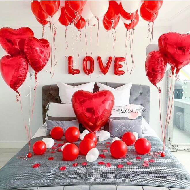 A bedroom decorated for Valentine's Day with red and white balloons, including heart shapes and a banner that reads 'Love'. The balloons are scattered around the room, some on the bed and others on the floor.