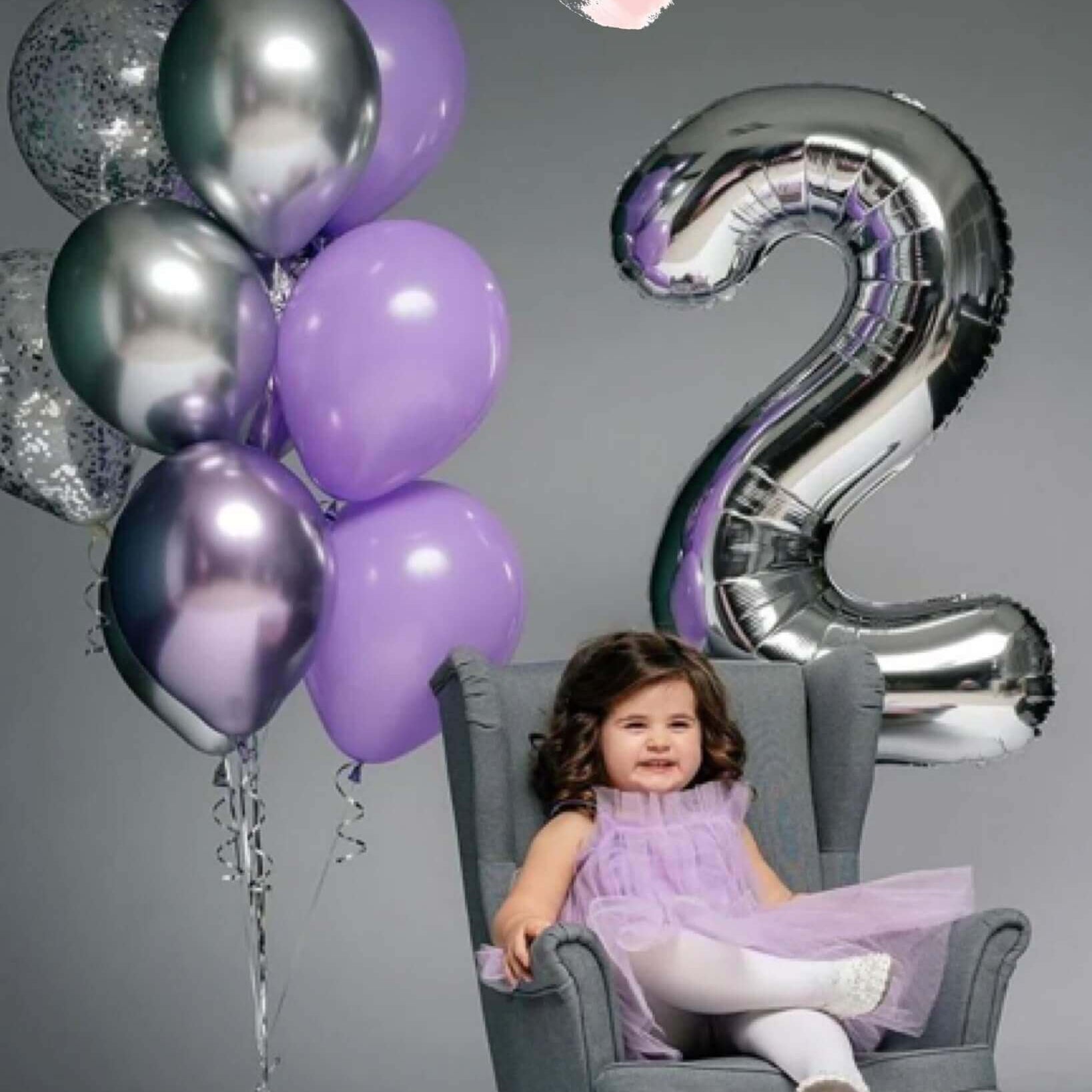 A child sitting in a chair with a '2' shaped balloon in front of them, with balloons in the background in a celebratory setting.