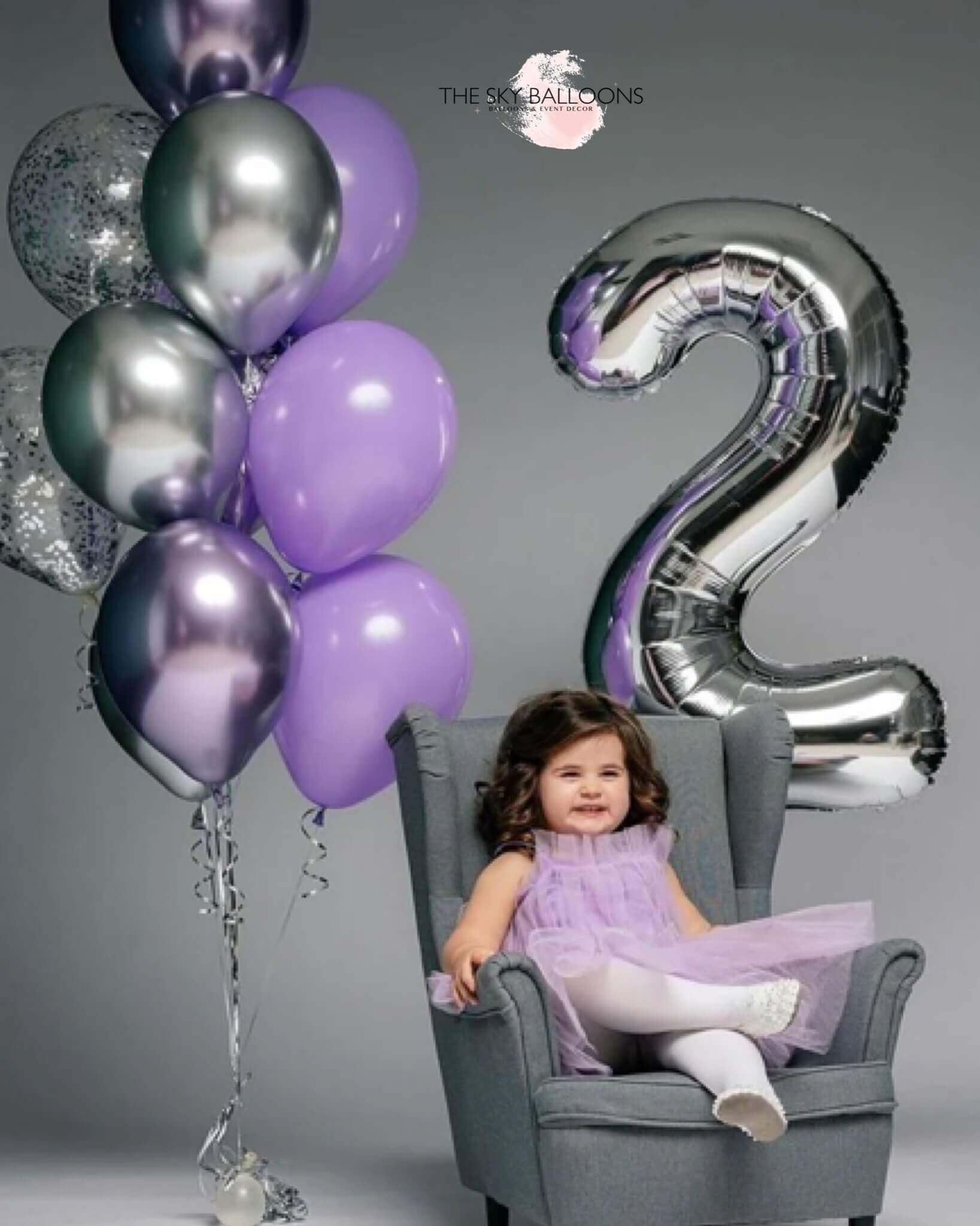 A child sitting in a chair with a '2' shaped balloon in front of them, with balloons in the background in a celebratory setting.