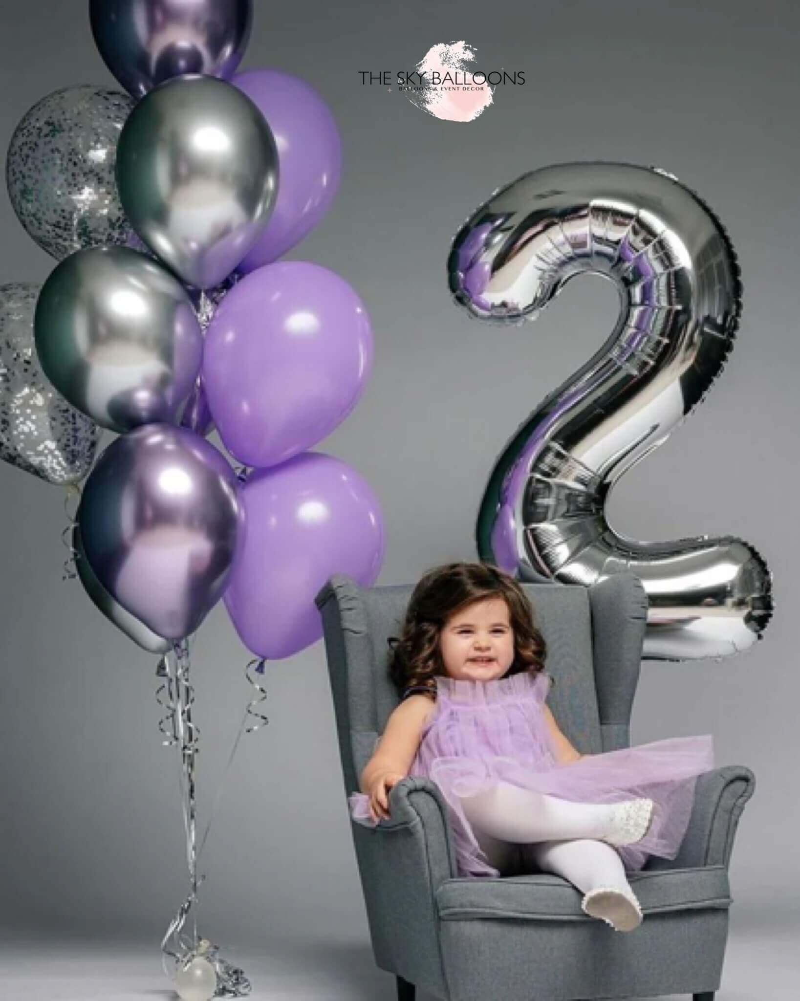 A child sitting in a chair with a '2' shaped balloon in front of them, with balloons in the background in a celebratory setting.
