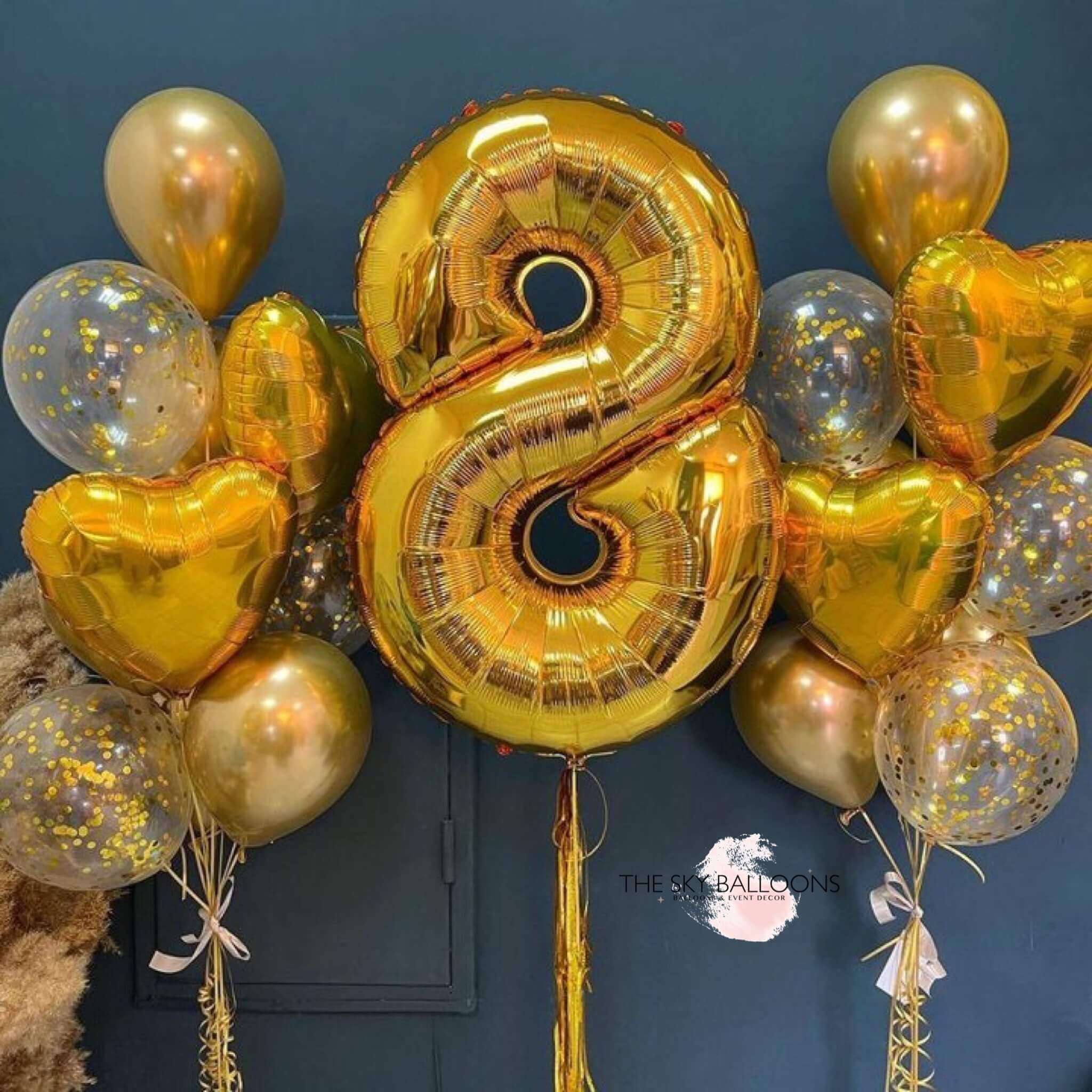 A bouquet of gold-colored balloons including a large number balloon, heart-shaped foils, and confetti balloons, tied together with ribbons.