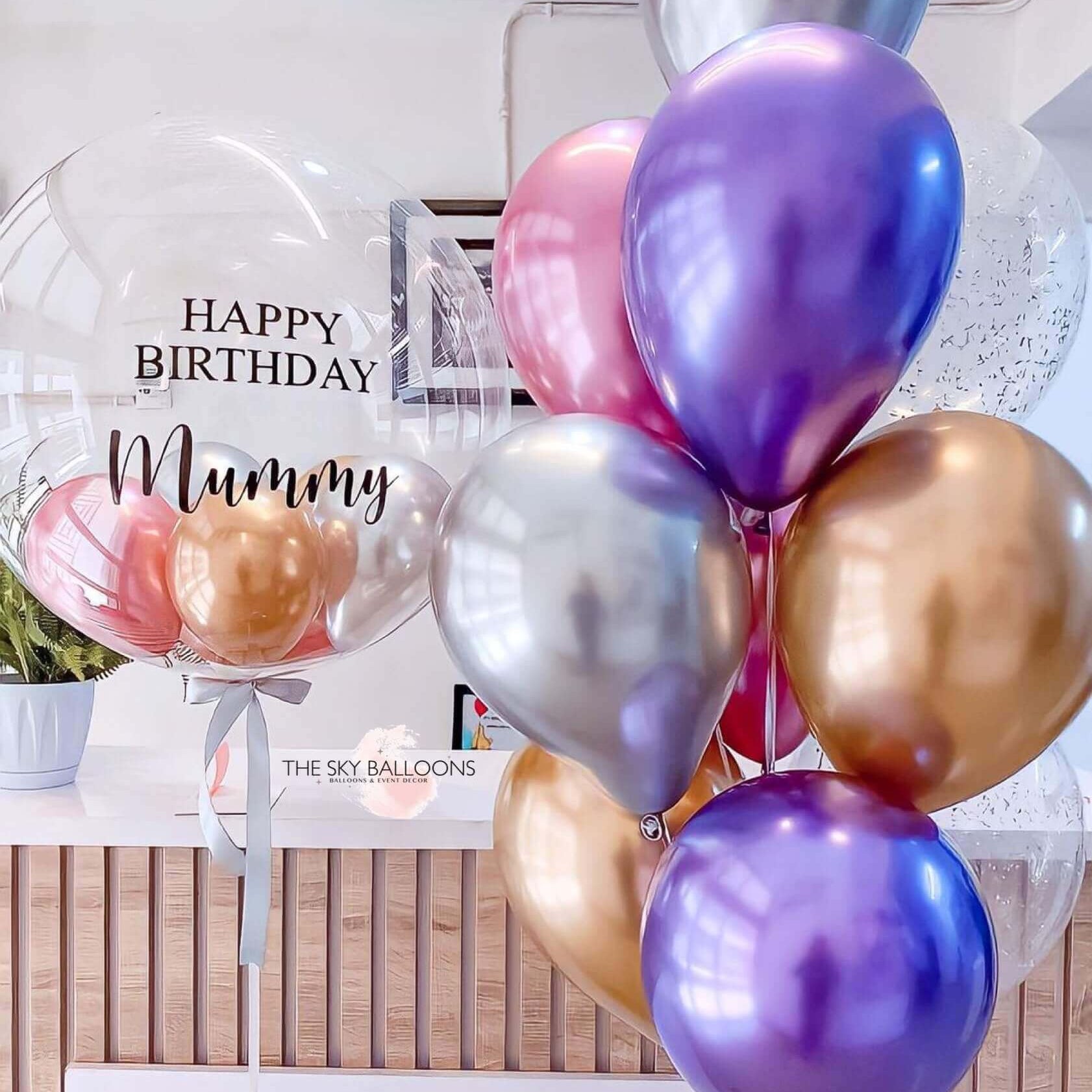 A bunch of helium-filled balloons including standard latex balloons in various sizes and a large customized balloon with 'Happy Mother's Day' text in gold and purple colors.