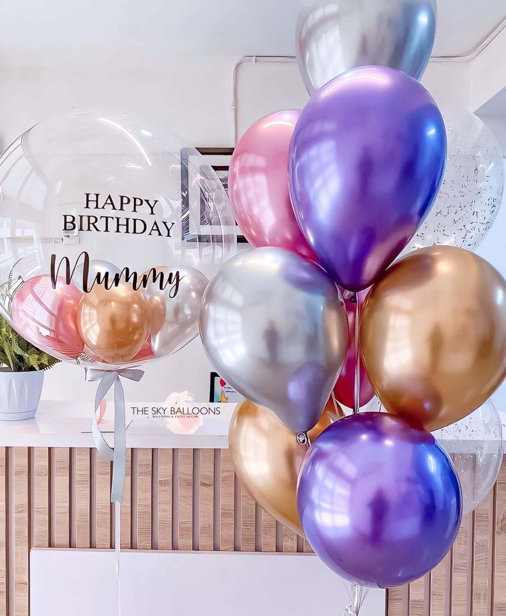 A bunch of helium-filled balloons including standard latex balloons in various sizes and a large customized balloon with 'Happy Mother's Day' text in gold and purple colors.