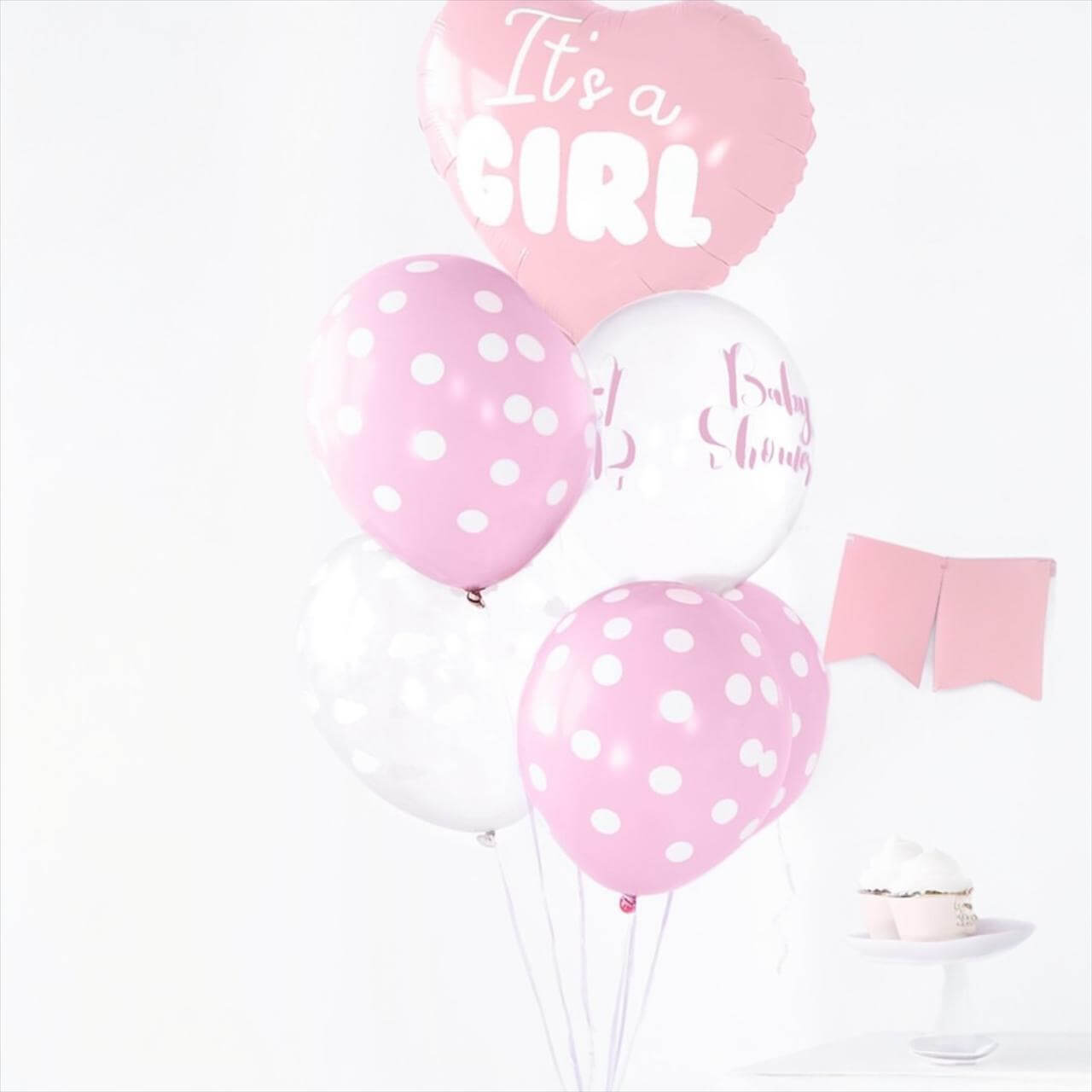 Pregnant woman in a pink dress holding 'It's a Girl' balloons against a pink background