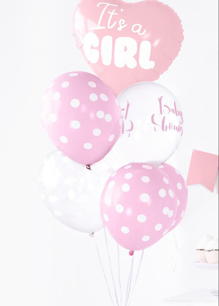 Pregnant woman in a pink dress holding 'It's a Girl' balloons against a pink background