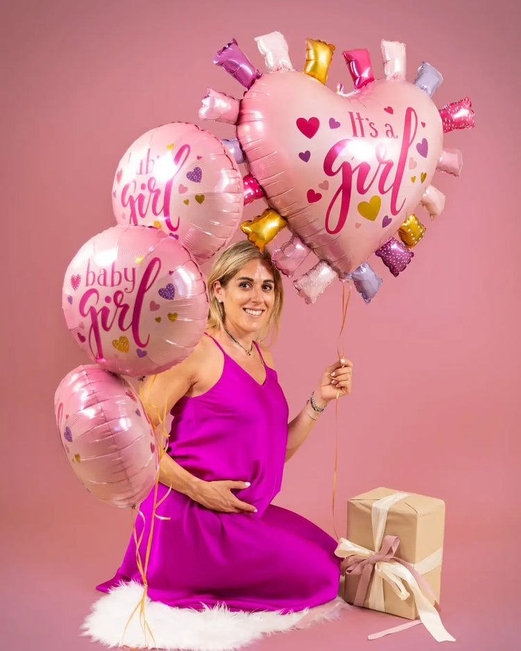 Pregnant woman in a pink dress holding 'It's a Girl' balloons against a pink background
