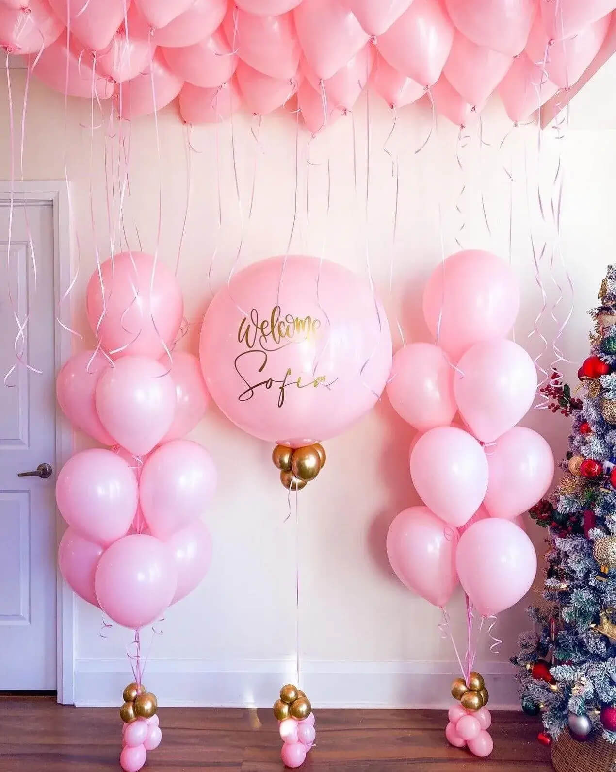 Pink balloon arch with 'Welcome Sofia' text, Christmas tree on right