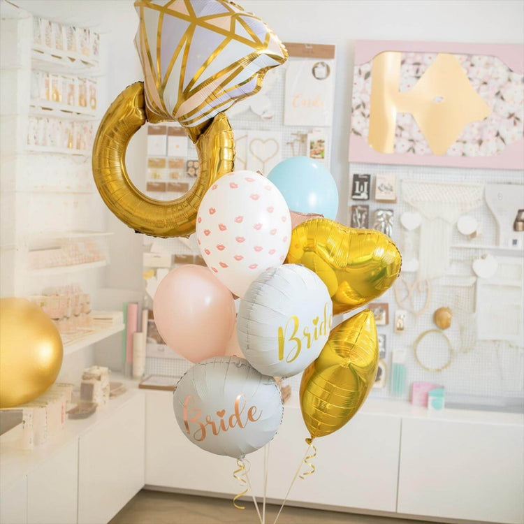 Three women in robes with 'Bride to be' balloons and a large silver ring on a pink background