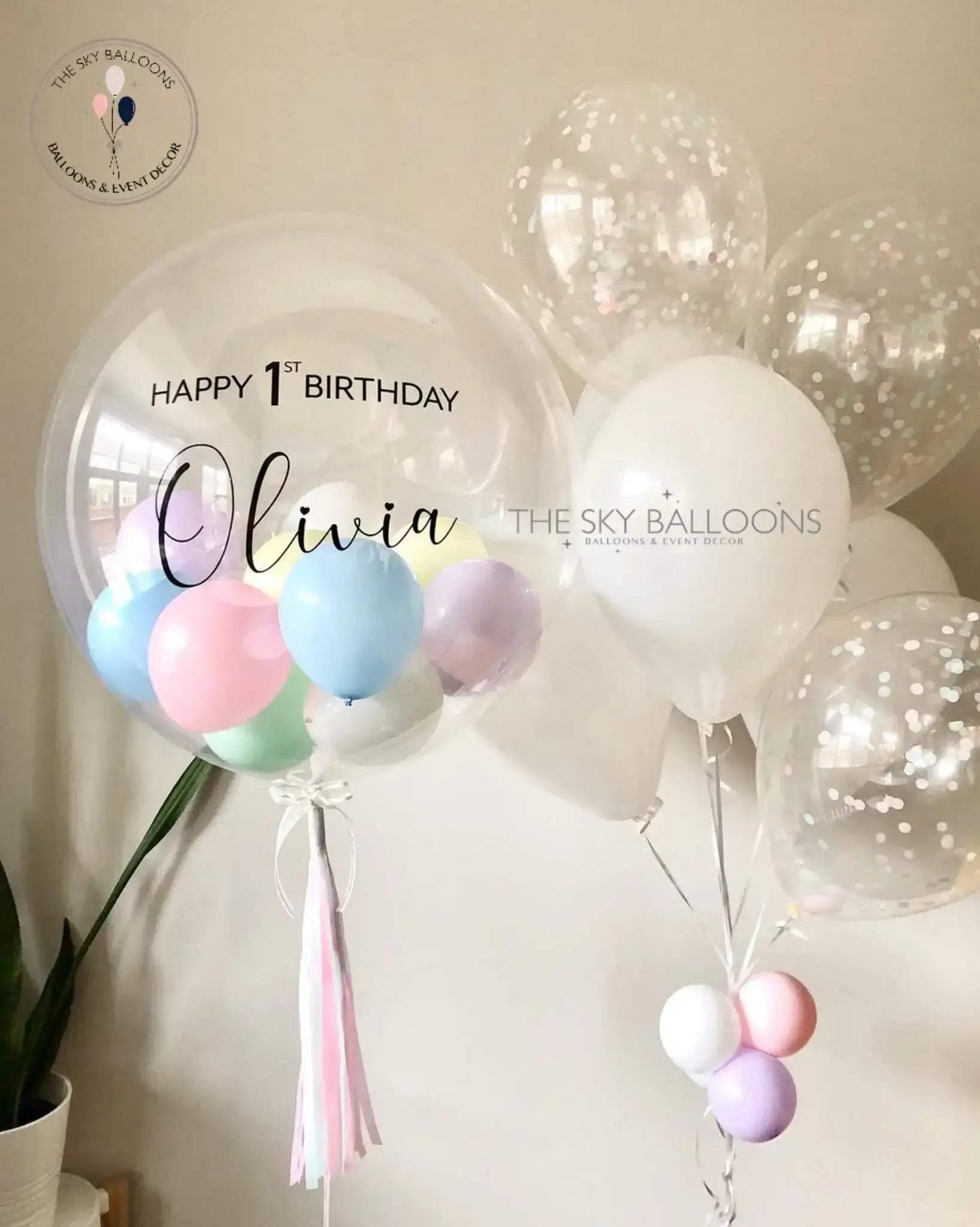 A balloon bouquet featuring a mix of round and oval-shaped balloons in shades of pink, white, blue, and green, with 'Happy 1st Birthday' and a name 'Olivia' printed on a pink banner.
