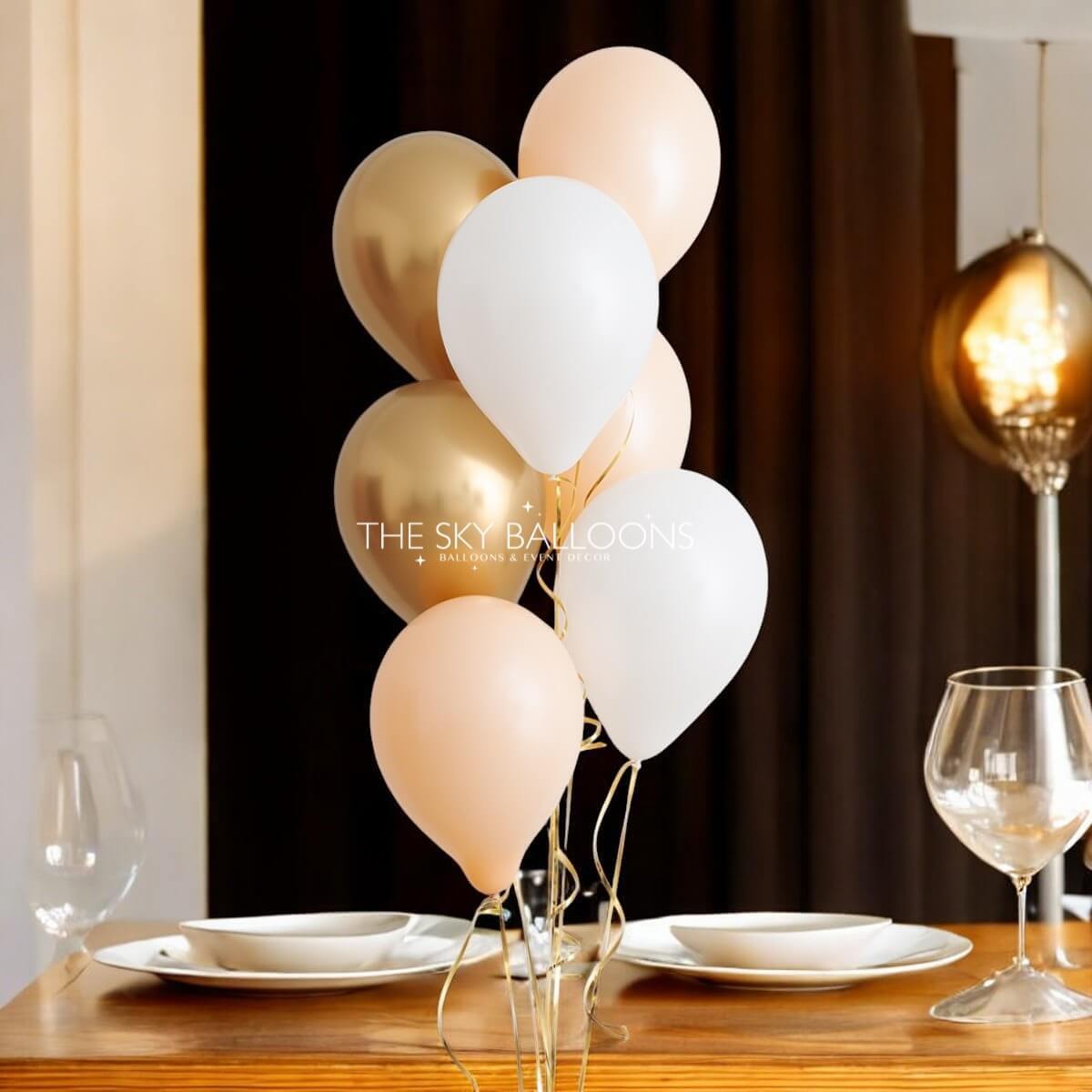 A bouquet of helium-filled balloons in various colors, including gold, white, and pink, tied together and placed on a table.