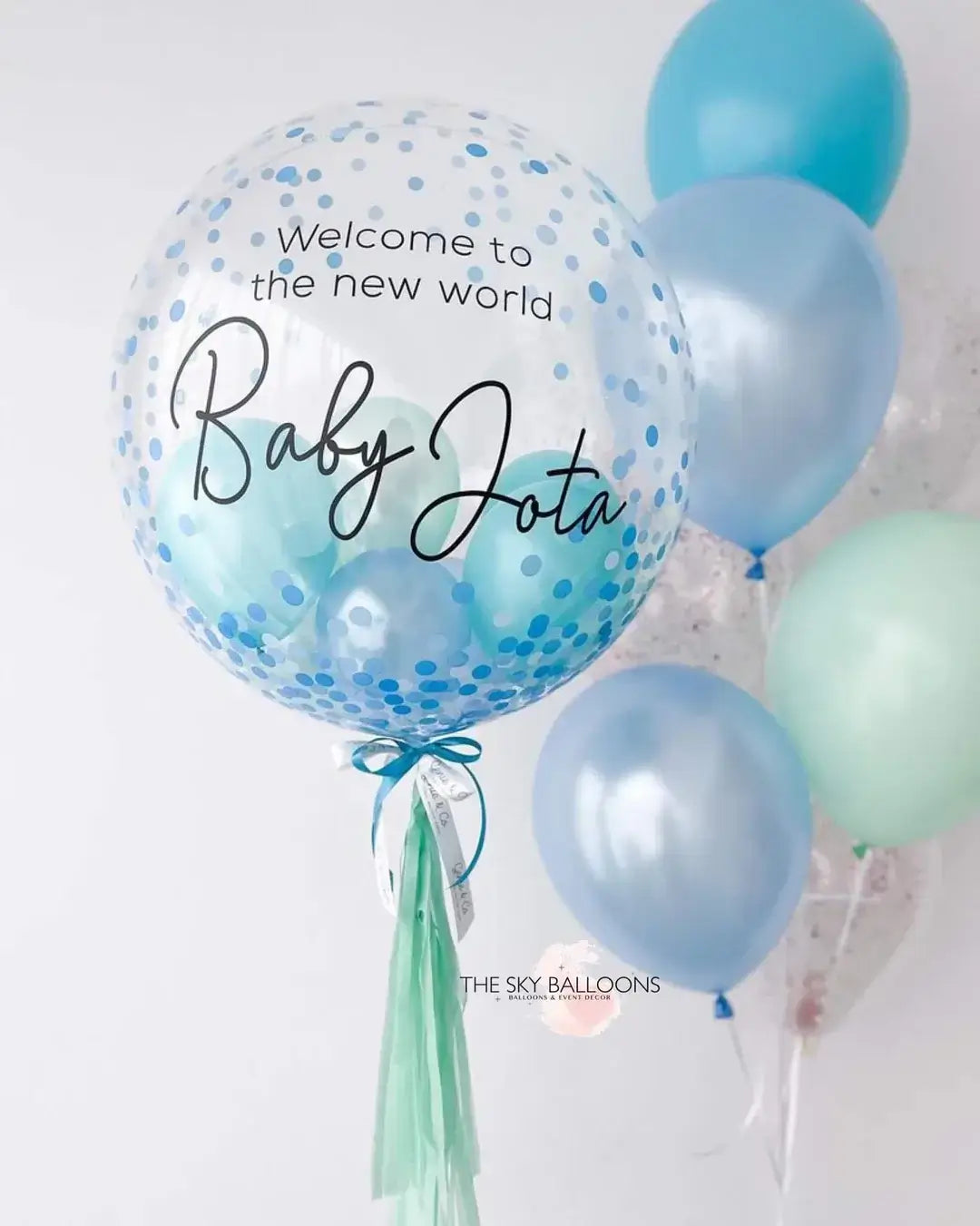 A bouquet of balloons featuring a large blue balloon with text, accompanied by smaller balloons in blue and white colors, with a tassel detail.