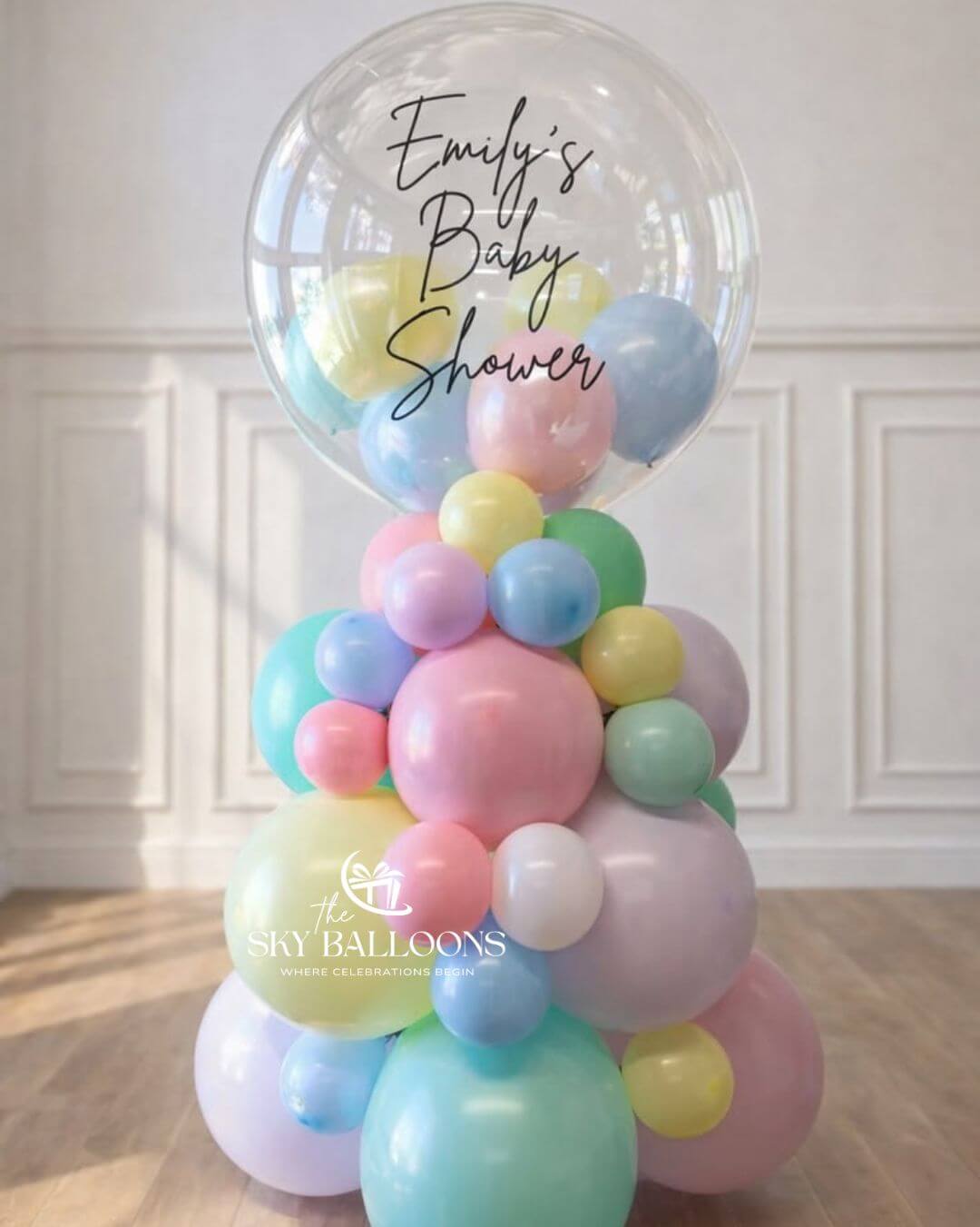 A pastel balloon garland balloon arch decoration for a baby shower, featuring a large clear balloon at the top with the text 'Emily's Baby Shower' and a cluster of pastel balloons in pink, blue, yellow, and green below. The balloons are arranged on a wooden floor against a white paneled wall.