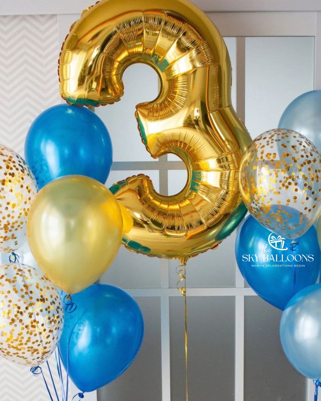 A festive balloon arrangement featuring a large metallic gold foil number 3 balloon, surrounded by blue, light blue, and clear balloons with gold confetti, set against a white paneled wall.