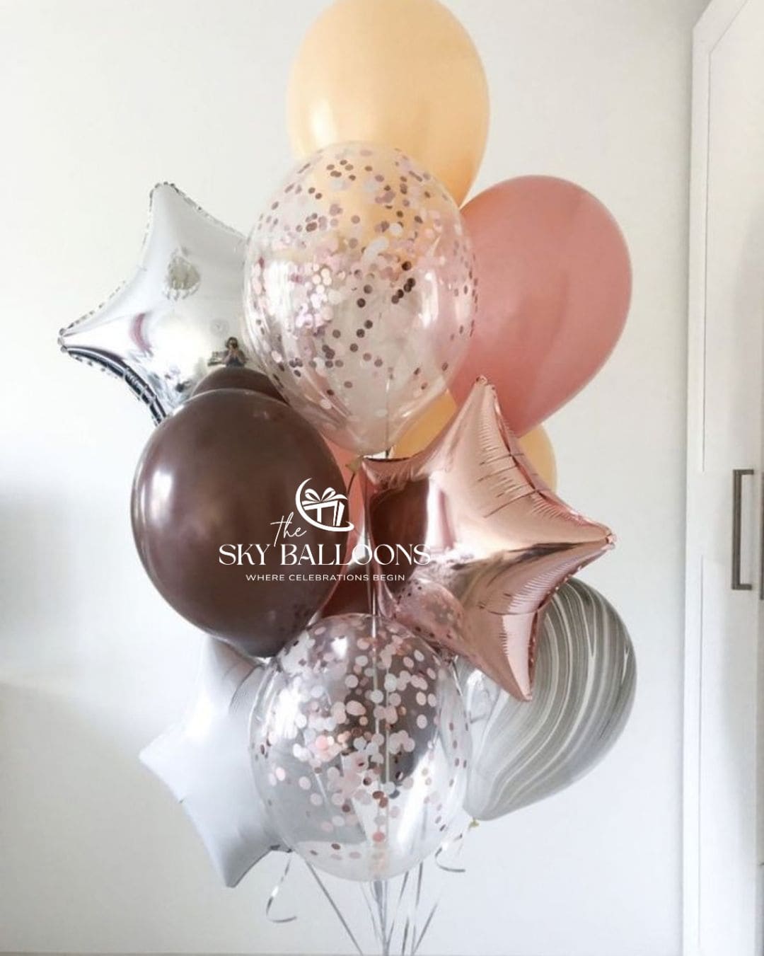 A cluster of decorative balloons including metallic rose gold star balloons, a metallic silver star balloon, a metallic dark brown round balloon, and several clear balloons filled with rose gold and silver confetti. 