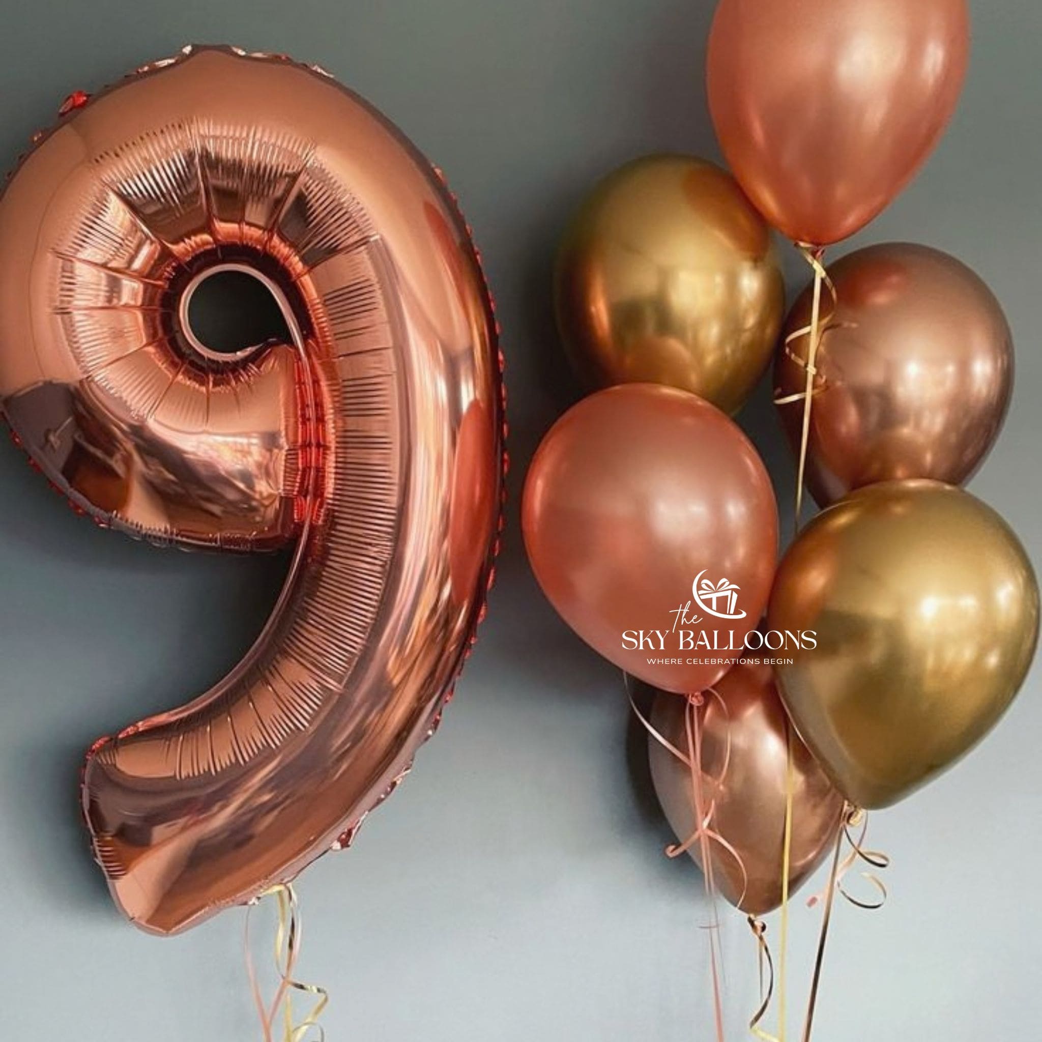 A metallic rose gold foil balloon shaped like the number 9, displayed next to a cluster of metallic balloons in rose gold and gold, against a dark gray background. 