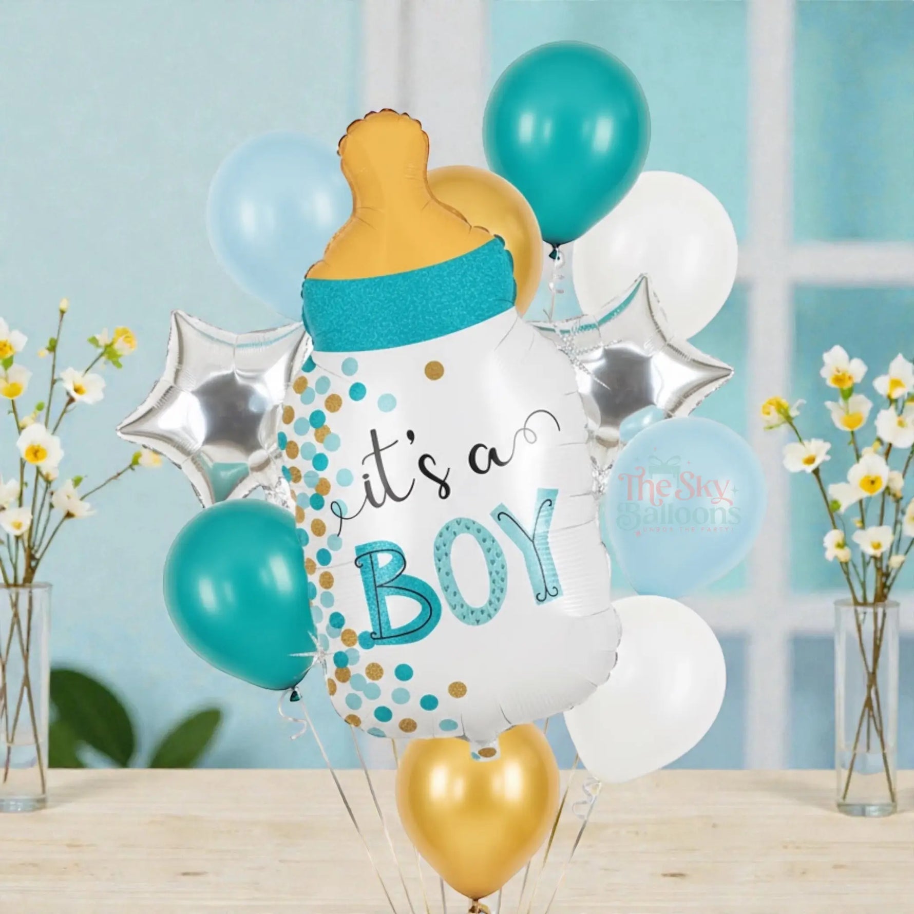 Balloon bouquet with 'it's a boy' message and baby bottle design on a table with flowers in the background.