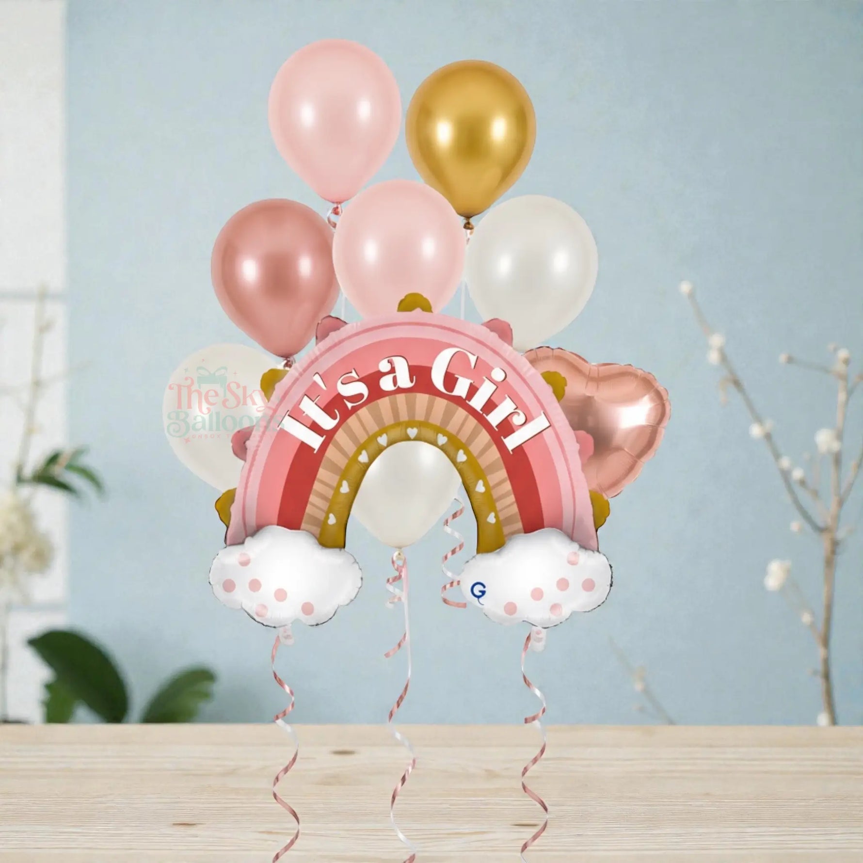 Balloon bouquet with rainbow and 'It's a Girl' text against a light blue wall.