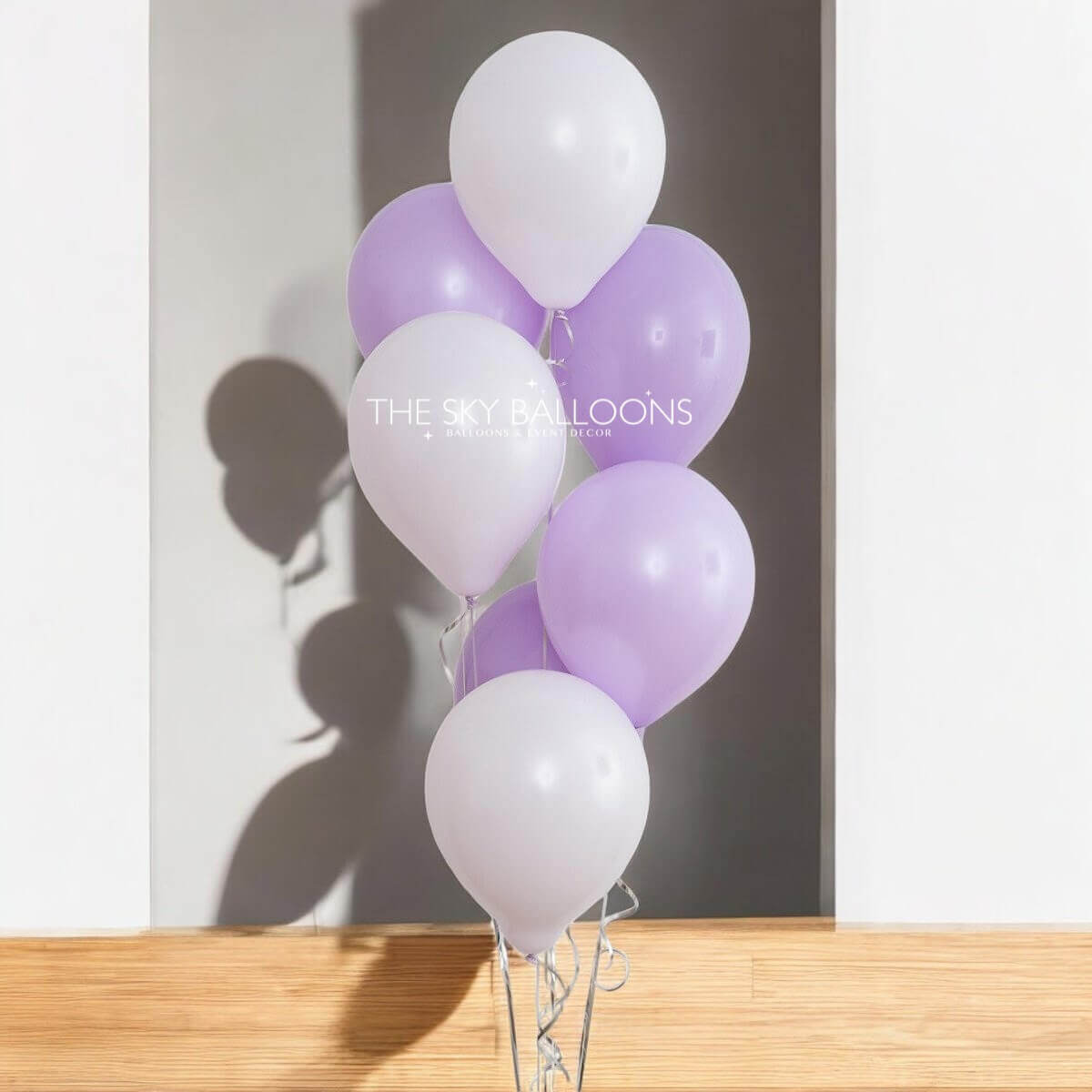 A bouquet of helium-filled purple and white balloons tied together, with the shadow of a person holding a camera reflected on the wall in the background.