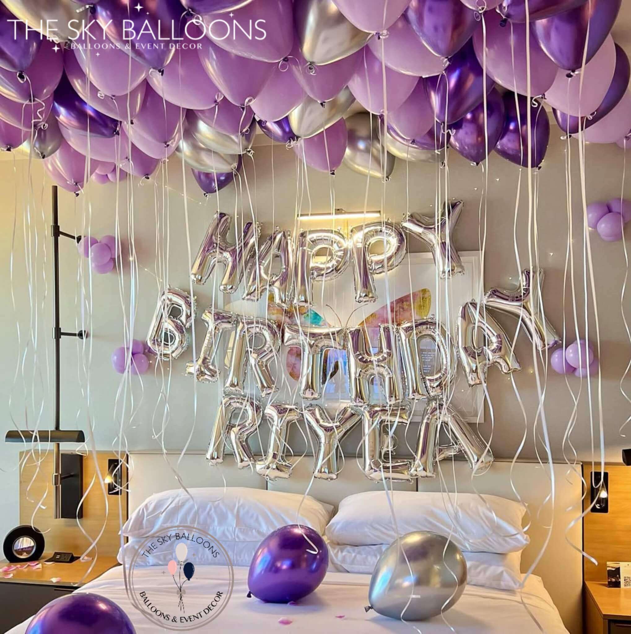 A room decorated with purple and silver helium balloons, featuring a happy birthday banner and personalized name.