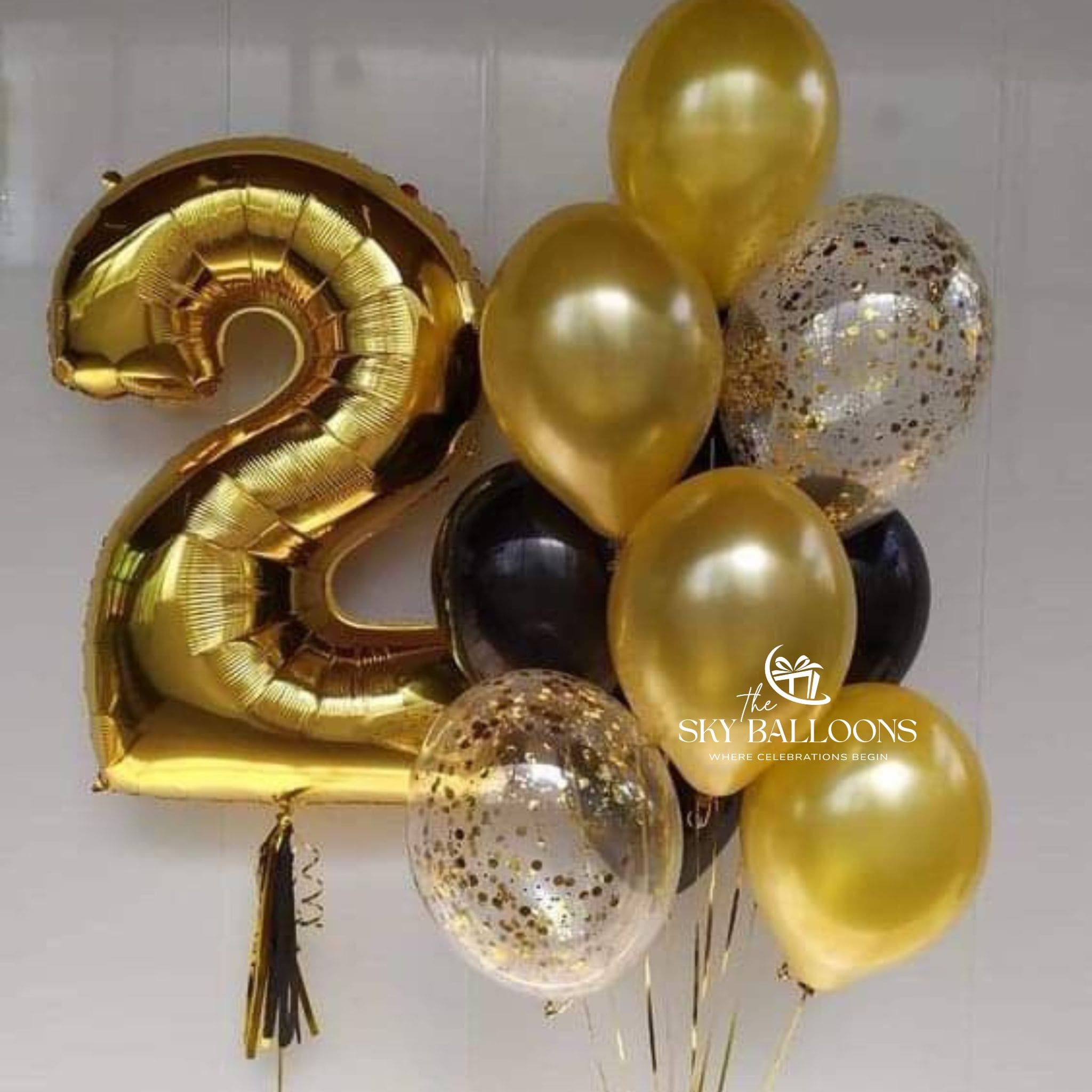 A balloon set featuring a large metallic gold foil balloon shaped like the number 2, surrounded by gold, black, and clear confetti balloons against a light gray background. A watermark for 'the SKY BALLOONS' is visible on the right side.