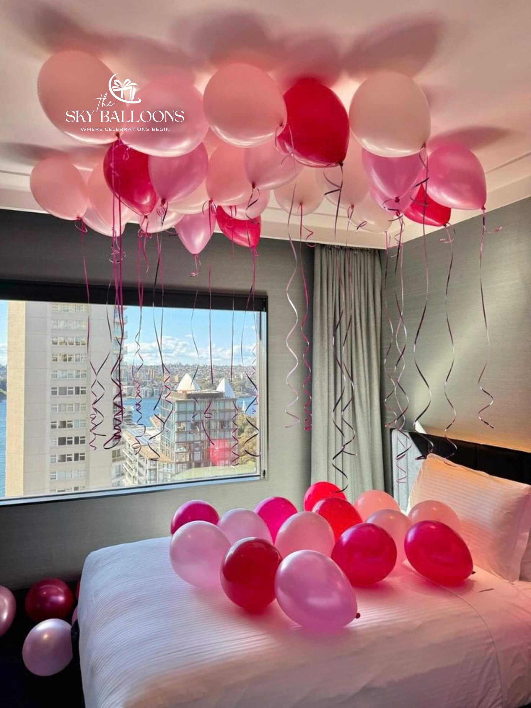 Pink and red balloon decoration in a bedroom, with a cluster of balloons floating near the ceiling and more balloons scattered on a white bed. 