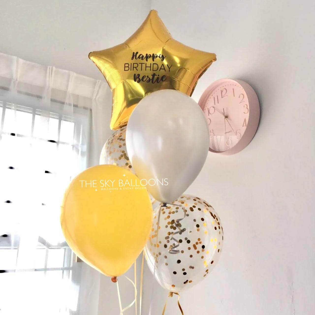 Balloons with 'Happy Birthday Bestie' on a wall with a clock.