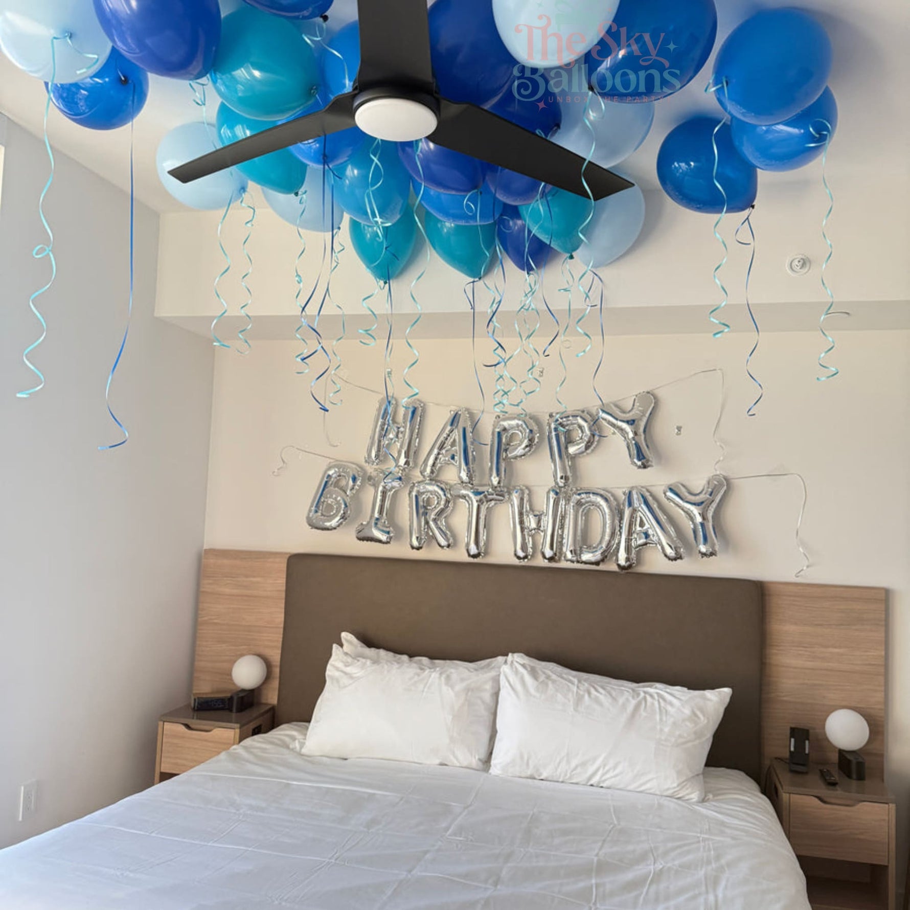 Bedroom with blue and white balloons, 'Happy Birthday' sign, and ceiling fan.