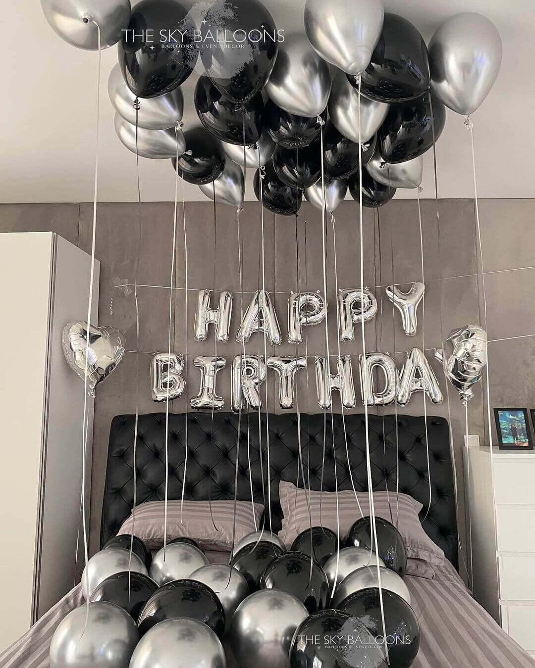 A bedroom setup with silver and black balloons, including a 'HAPPY BIRTHDAY' banner, ceiling balloons, and heart-shaped foil balloons.