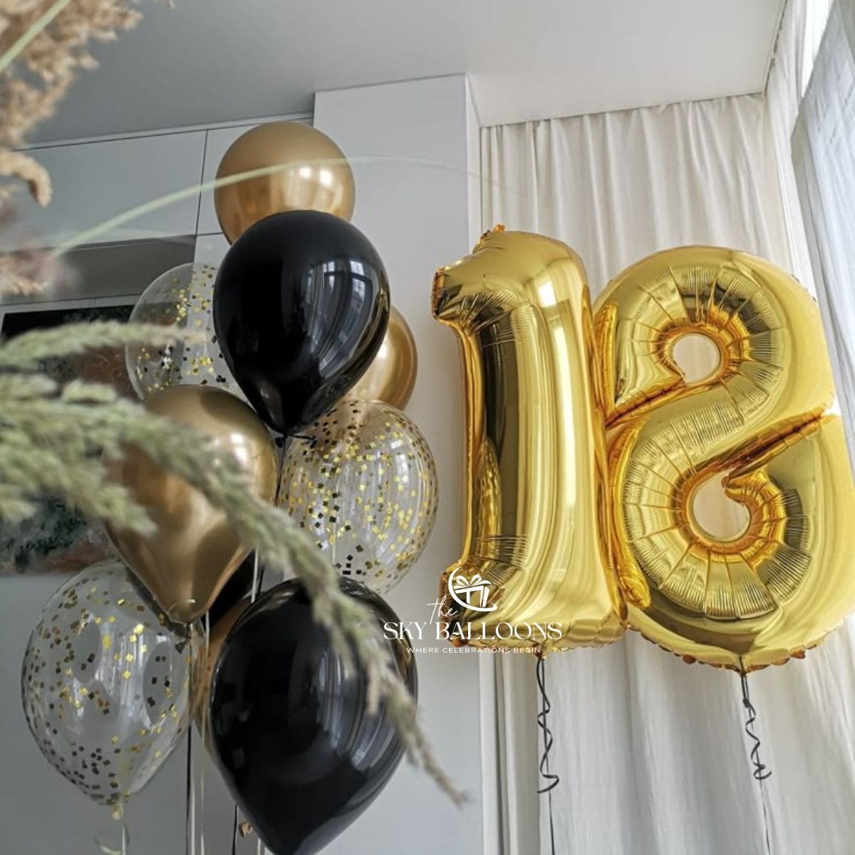 Gold and black foil balloons, including a large number 18 balloon, displayed in a decorated room with white curtains and dried grasses.