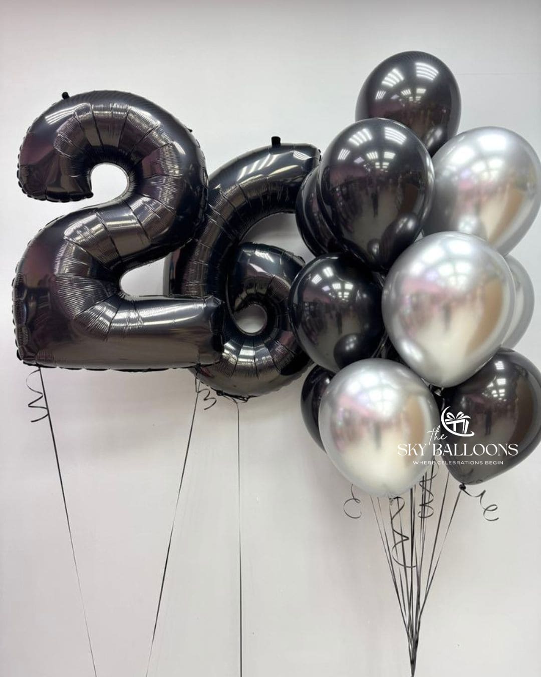 A set of metallic foil balloons including a large number '2' and '0' balloon, alongside a cluster of round balloons in black, silver, and dark gray, displayed against a plain light background. A watermark for 'The Sky Balloons' is visible on the right side.