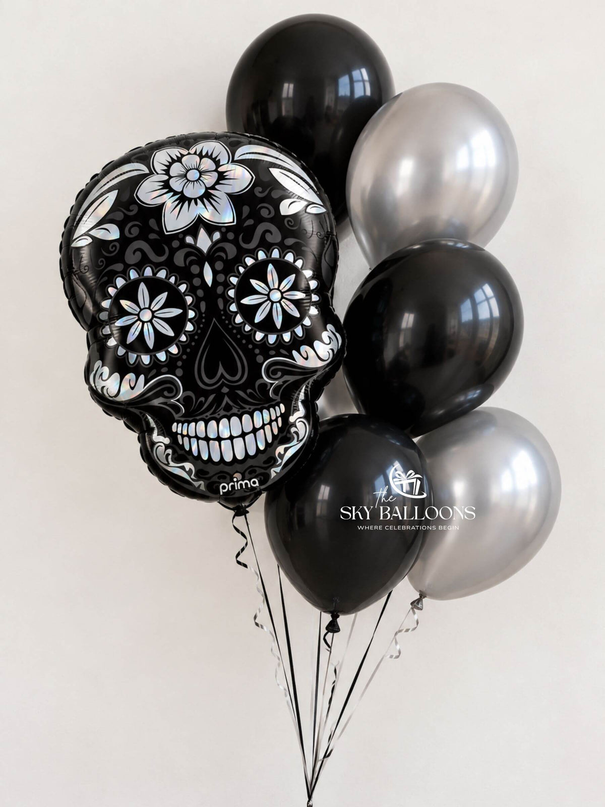 A cluster of decorative balloons including a large black foil balloon shaped like a sugar skull with white floral and ornamental details, alongside several matte black and shiny metallic silver balloons, tied together with black strings against a plain light background.