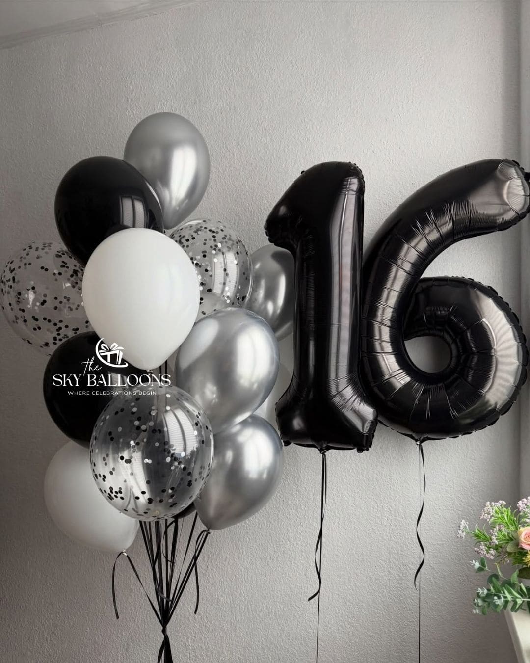 A cluster of birthday balloons including black, white, metallic silver, and clear balloons with black confetti, featuring a large black foil number 16 balloon, set against a light gray wall.