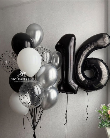 A cluster of birthday balloons including black, white, metallic silver, and clear balloons with black confetti, featuring a large black foil number 16 balloon, set against a light gray wall.