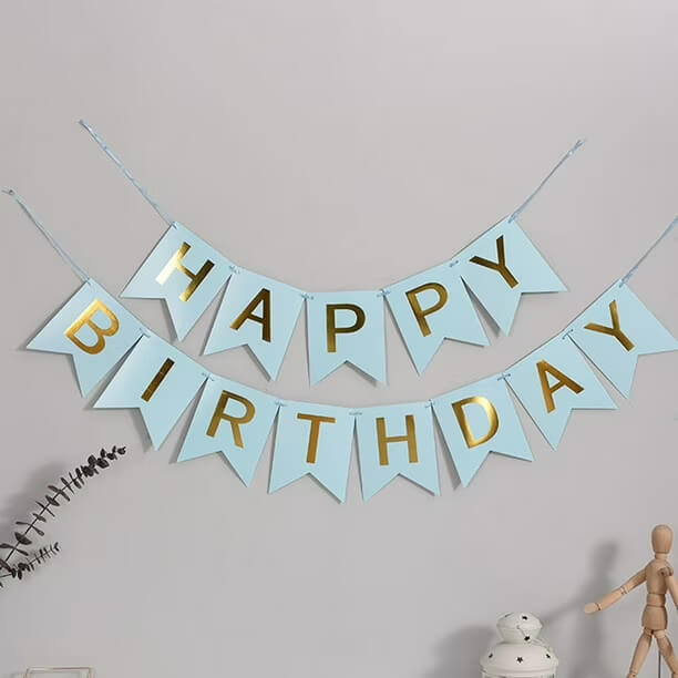 A light blue and gold 'HAPPY BIRTHDAY' banner garland hanging on a light gray wall, with small decorative props at the bottom corners.