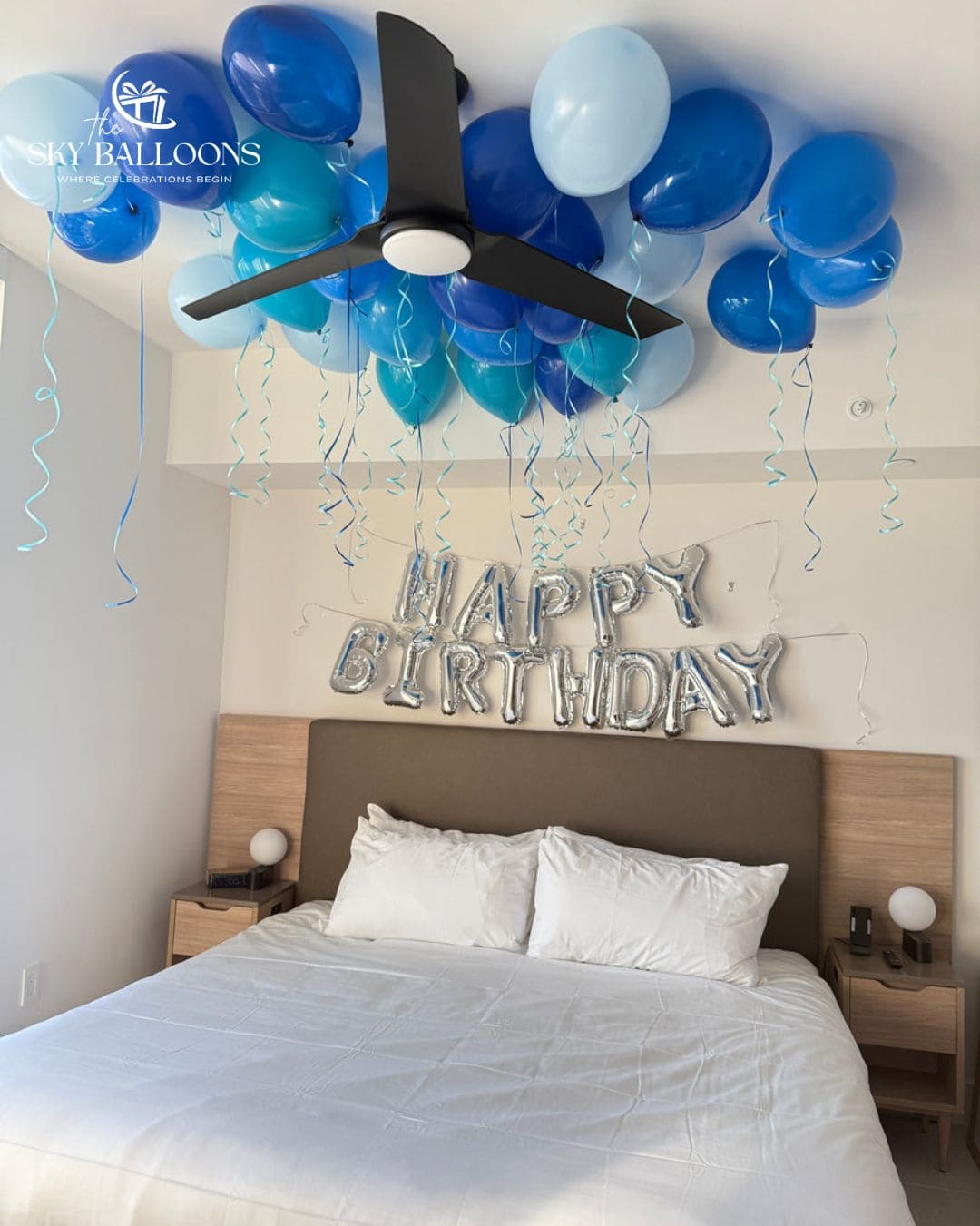 Birthday bedroom decoration with silver “Happy Birthday” letter balloons above the bed and dark blue, light blue and turquoise ceiling balloons with curling ribbons.⠋