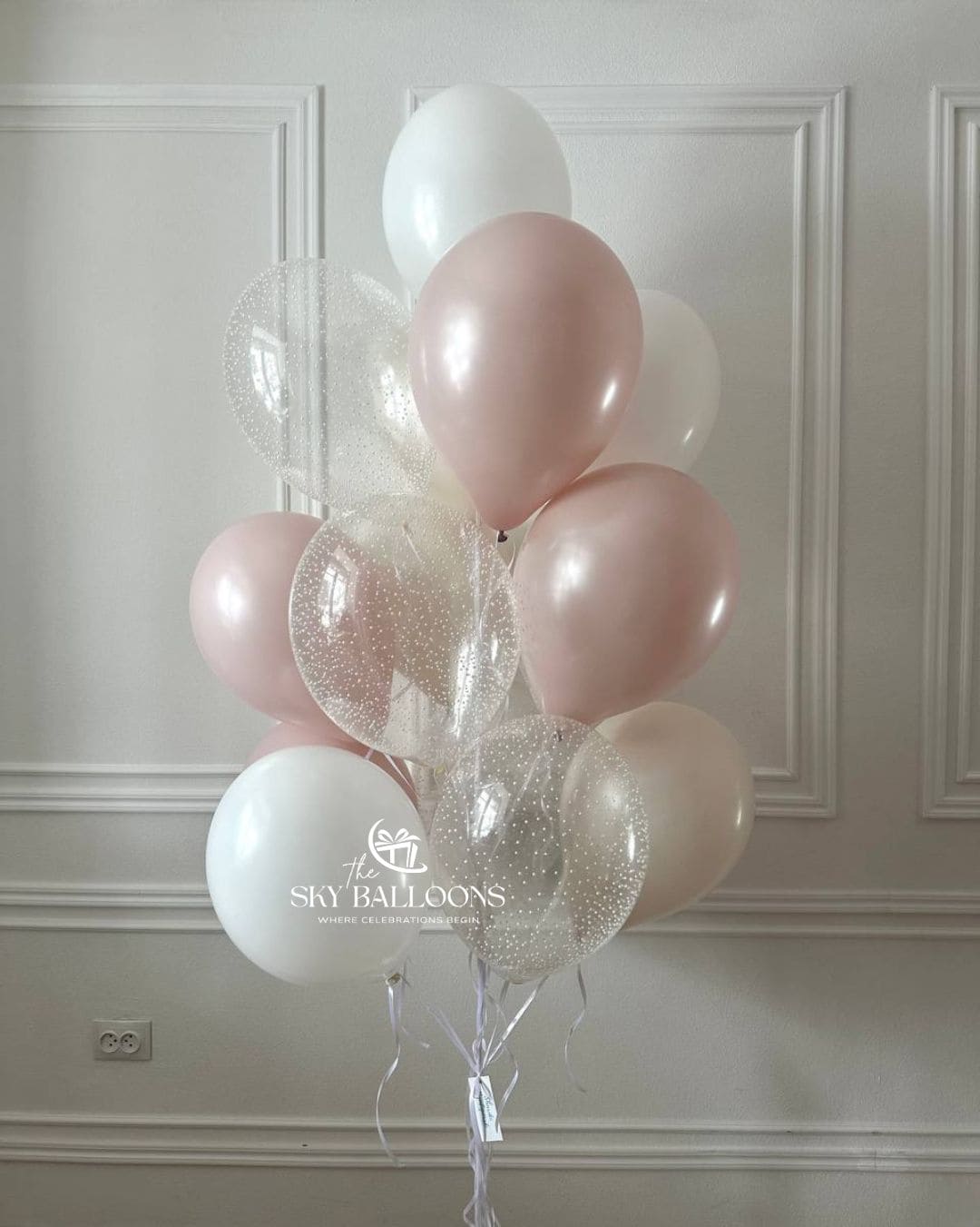 Balloon bouquet with blush pink and white latex balloons and clear glitter-filled balloons, tied with white ribbons against a white panelled wall.