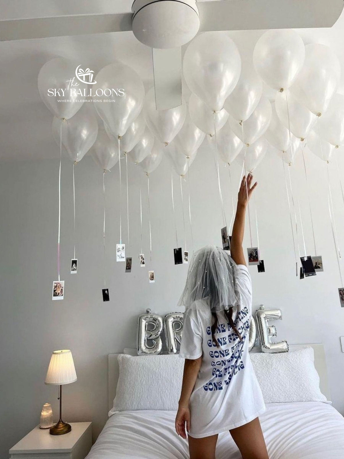 A woman stands on a bed in a bedroom, reaching toward a large balloon garland made of clear balloons suspended from the ceiling. Small photo prints hang from the balloons, and silver letter balloons spell 'BRIDE' behind her.